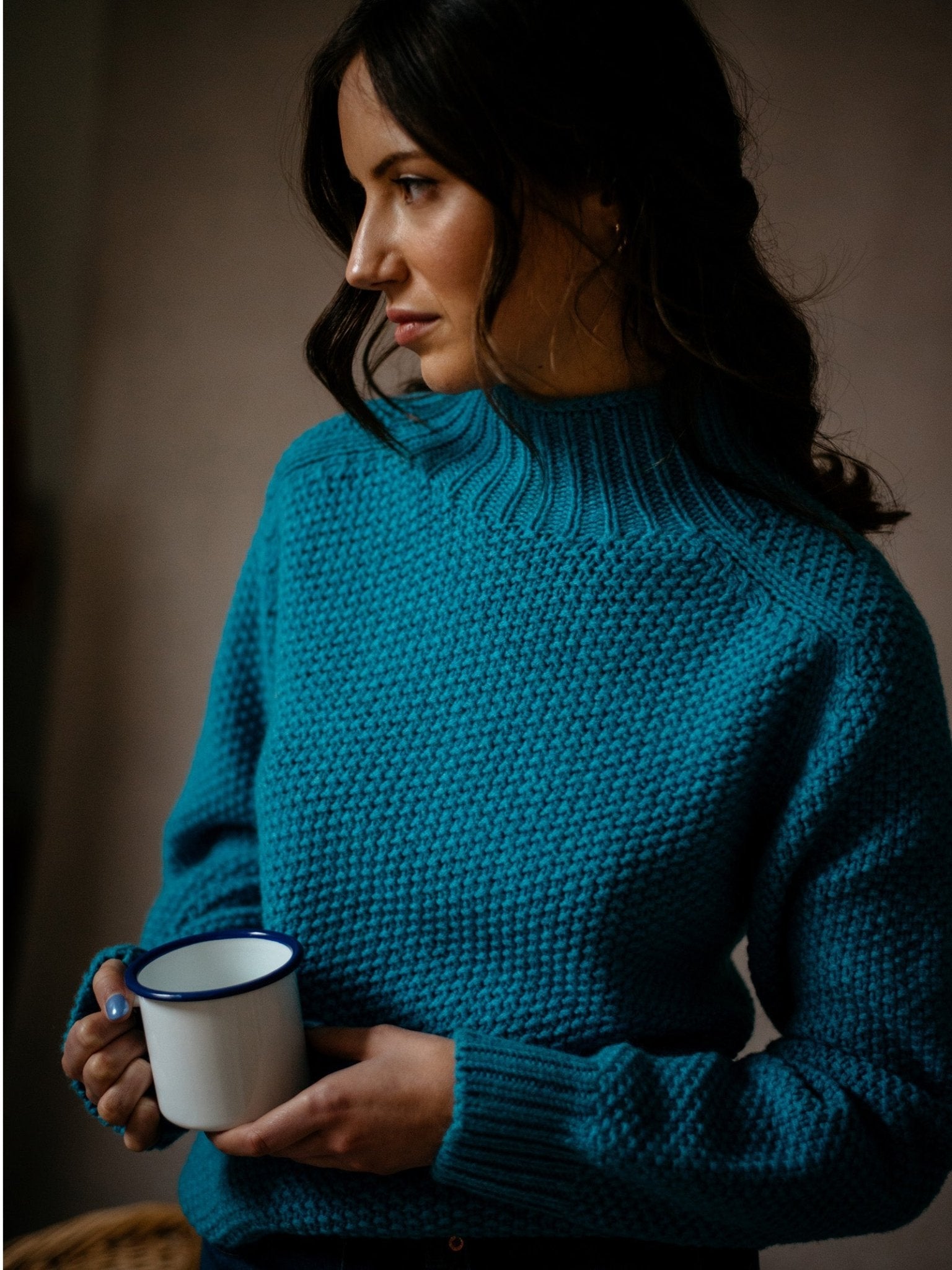 A woman with wavy dark hair, wearing a Chunky Moss Stitch Polo by Campbells of Beauly, holds a white mug with a blue rim and gazes thoughtfully to the side.
