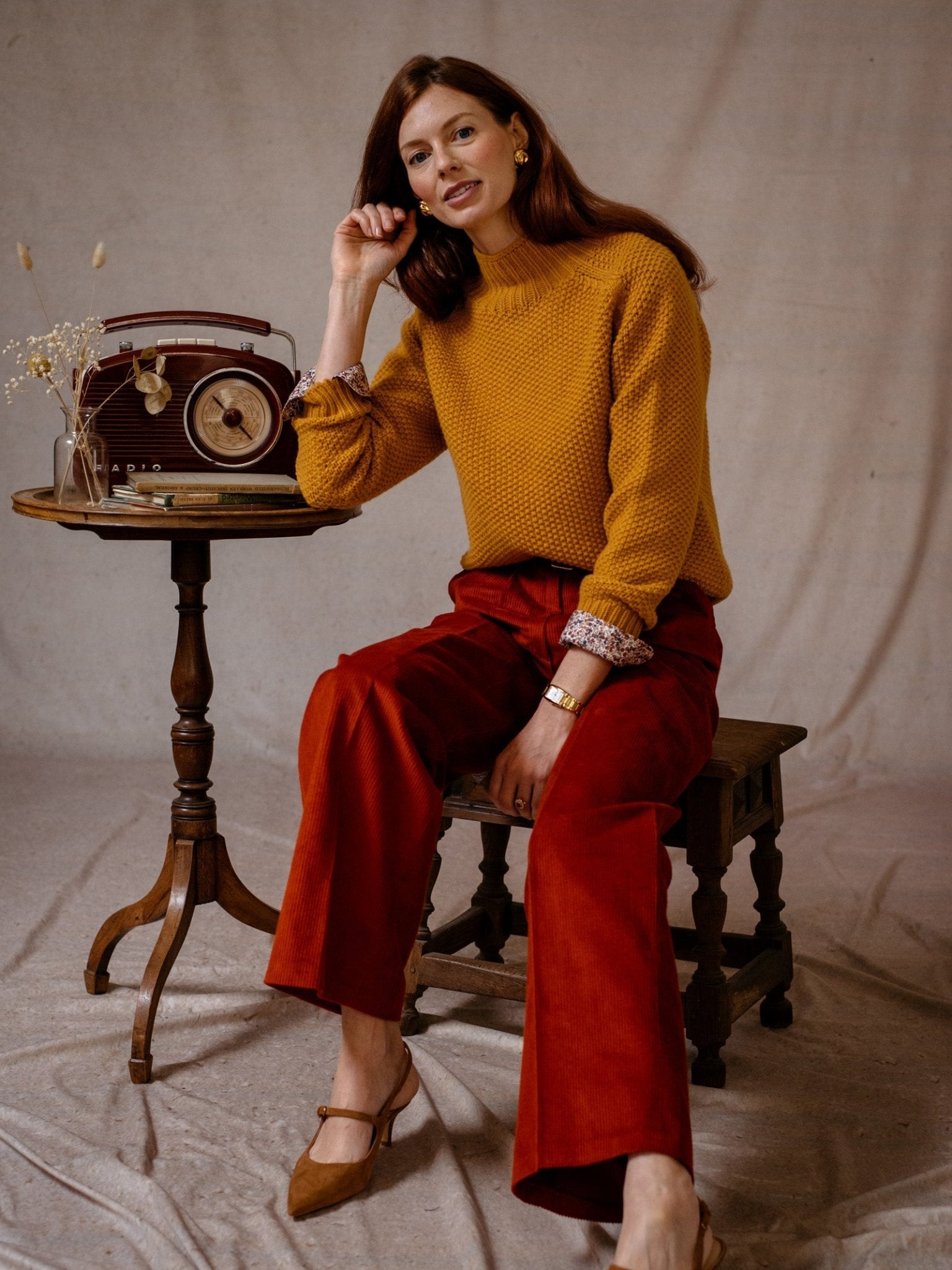 A woman with long red hair sits on a wooden stool, wearing the Campbells of Beauly Chunky Moss Stitch Polo in mustard yellow and red pants. She rests her elbow on a round table with a vintage radio, dried flowers, and books against a neutral fabric backdrop.