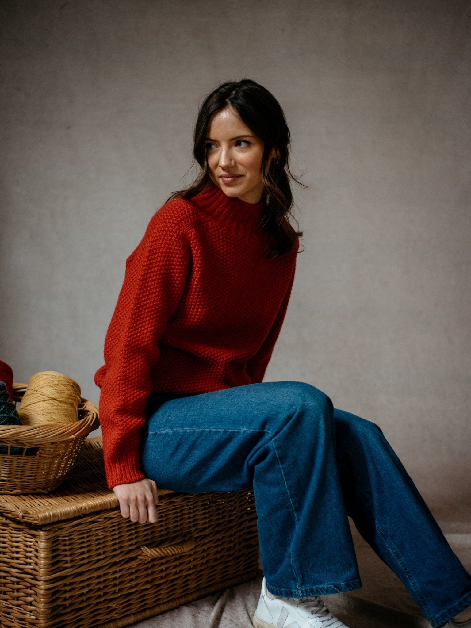 A woman in the Campbells of Beauly Chunky Moss Stitch Polo and blue jeans sits on a wicker chest with yarn beside her. She has shoulder-length brown hair, looks to the side with a slight smile, and is set against a plain, neutral background.