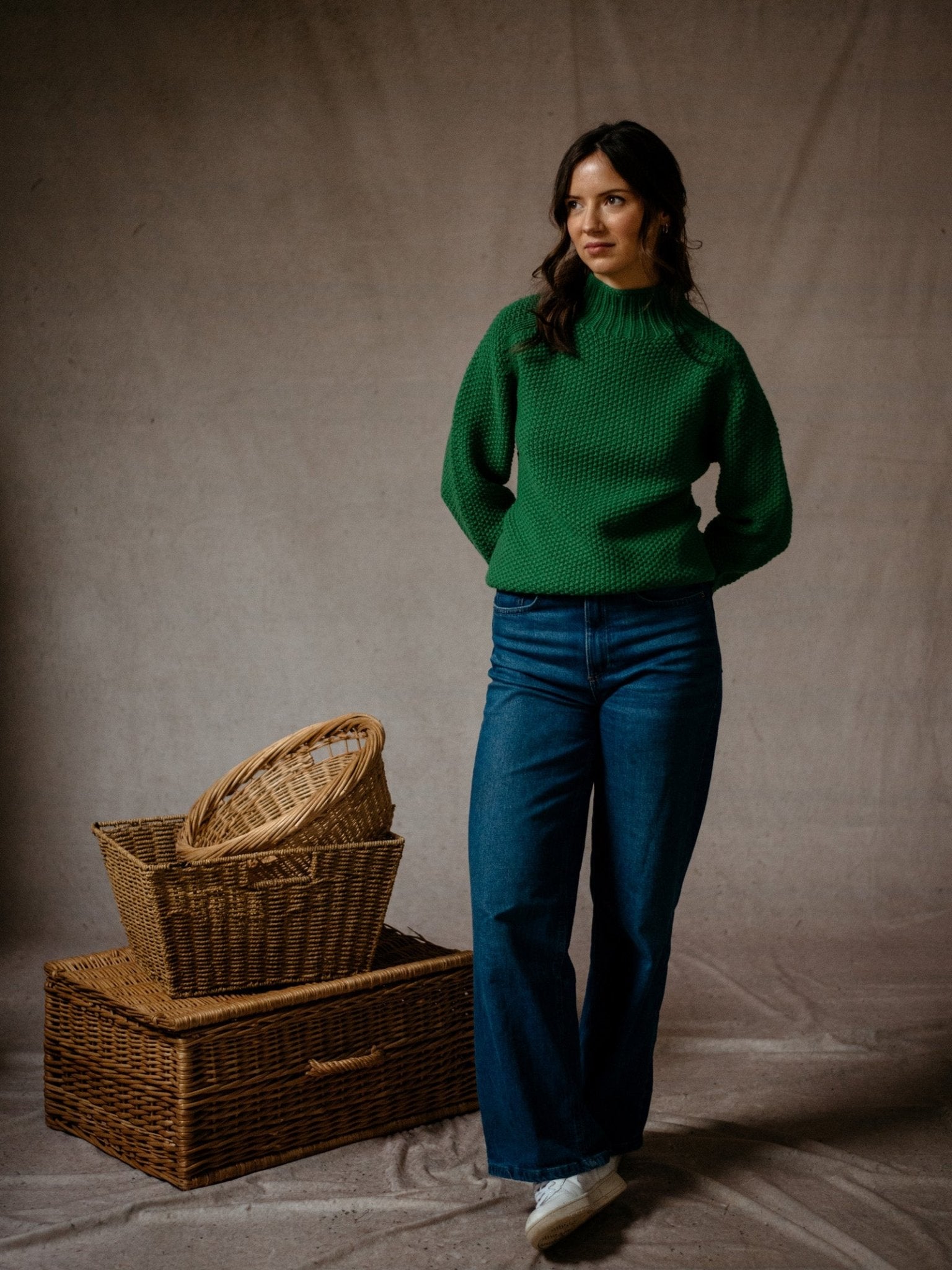 A woman in the Campbells of Beauly Chunky Moss Stitch Polo and blue jeans stands beside stacked wicker baskets against a neutral fabric backdrop, looking to the side with her hands behind her back.