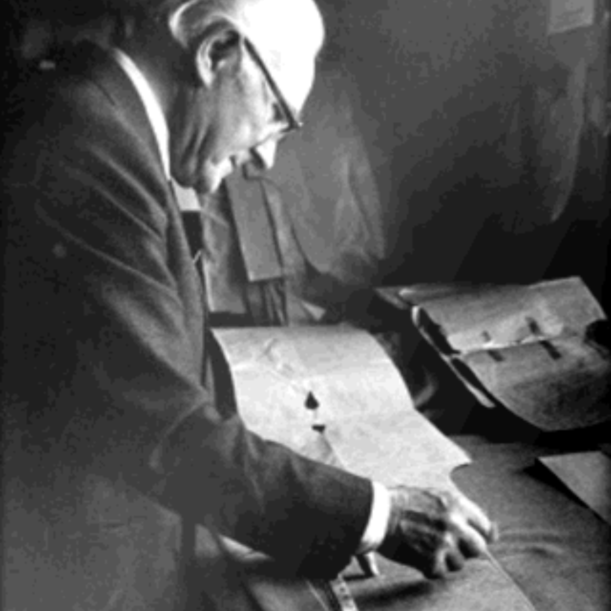 An elderly man in glasses and a suit examines large sheets of paper on a desk from Campbells of Beauly, appearing focused and intent on his work in a dimly lit room.