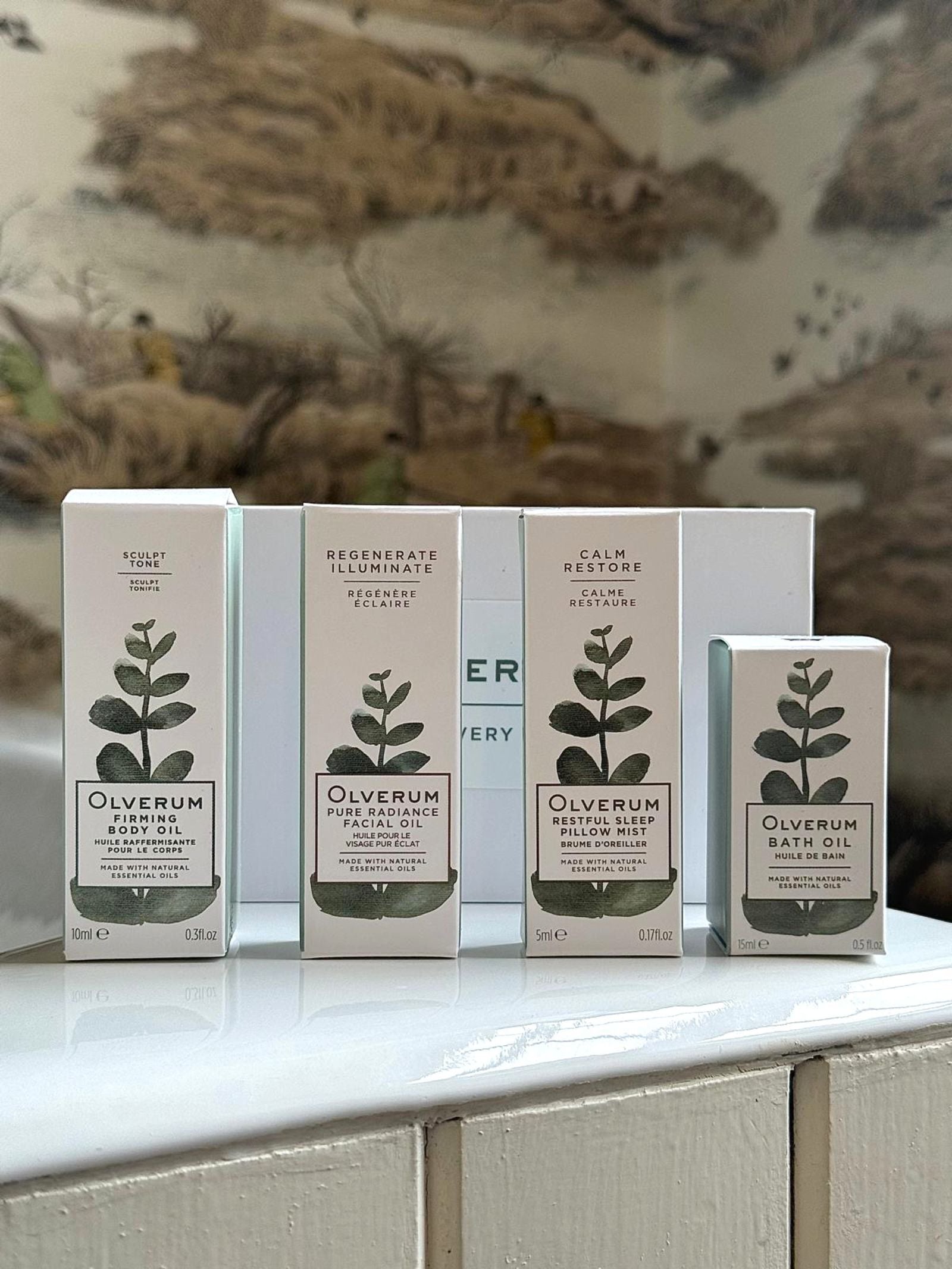 Four OLVERUM Discovery Box sets with minimalist, botanical packaging are arranged on a white surface. A blurred, nature-inspired background evokes the soothing essence of their renowned bath and body oils.