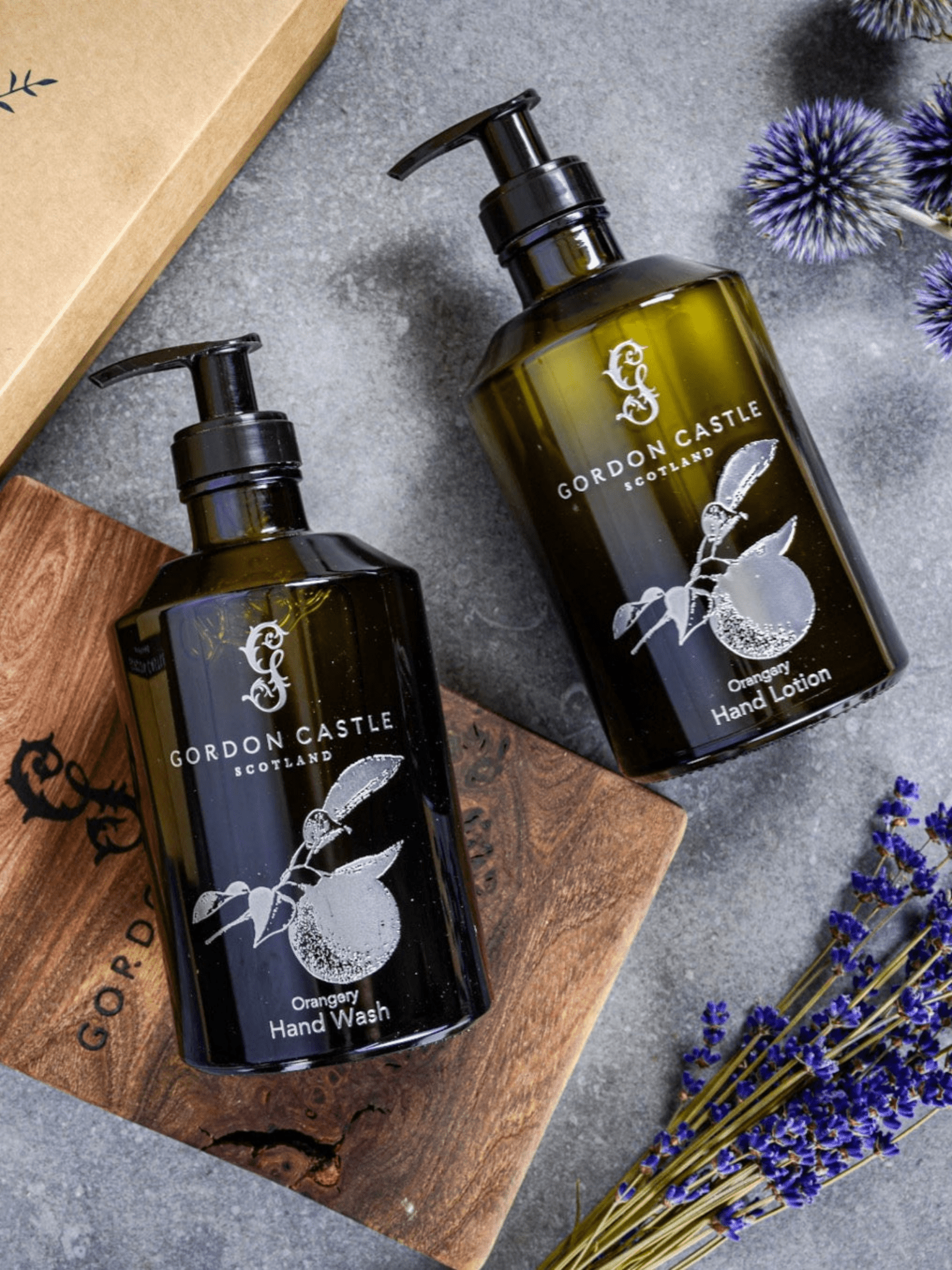 Two refillable Gordon Castle Orangery Hand Lotion glass bottles sit on a wooden board with lavender and thistle, highlighting sustainable luxury next to a cardboard box.
