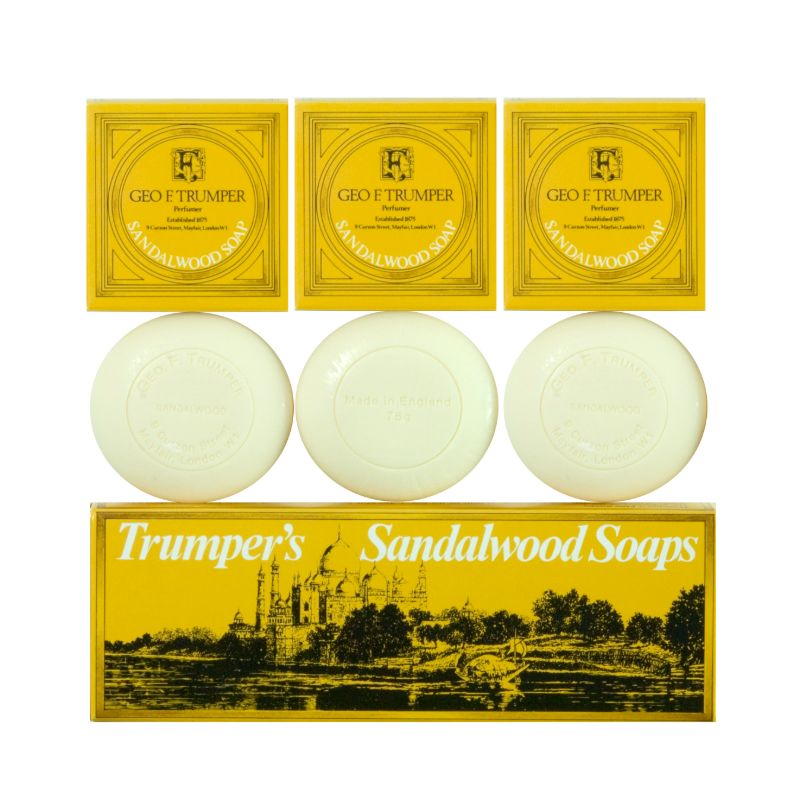Three oval white soaps from the Geo. F. Trumper Sandalwood Hand Soaps Set of 3, each in front of a yellow box with brand labeling and a larger box below reading Trumpers Sandalwood Soaps featuring a building illustration.