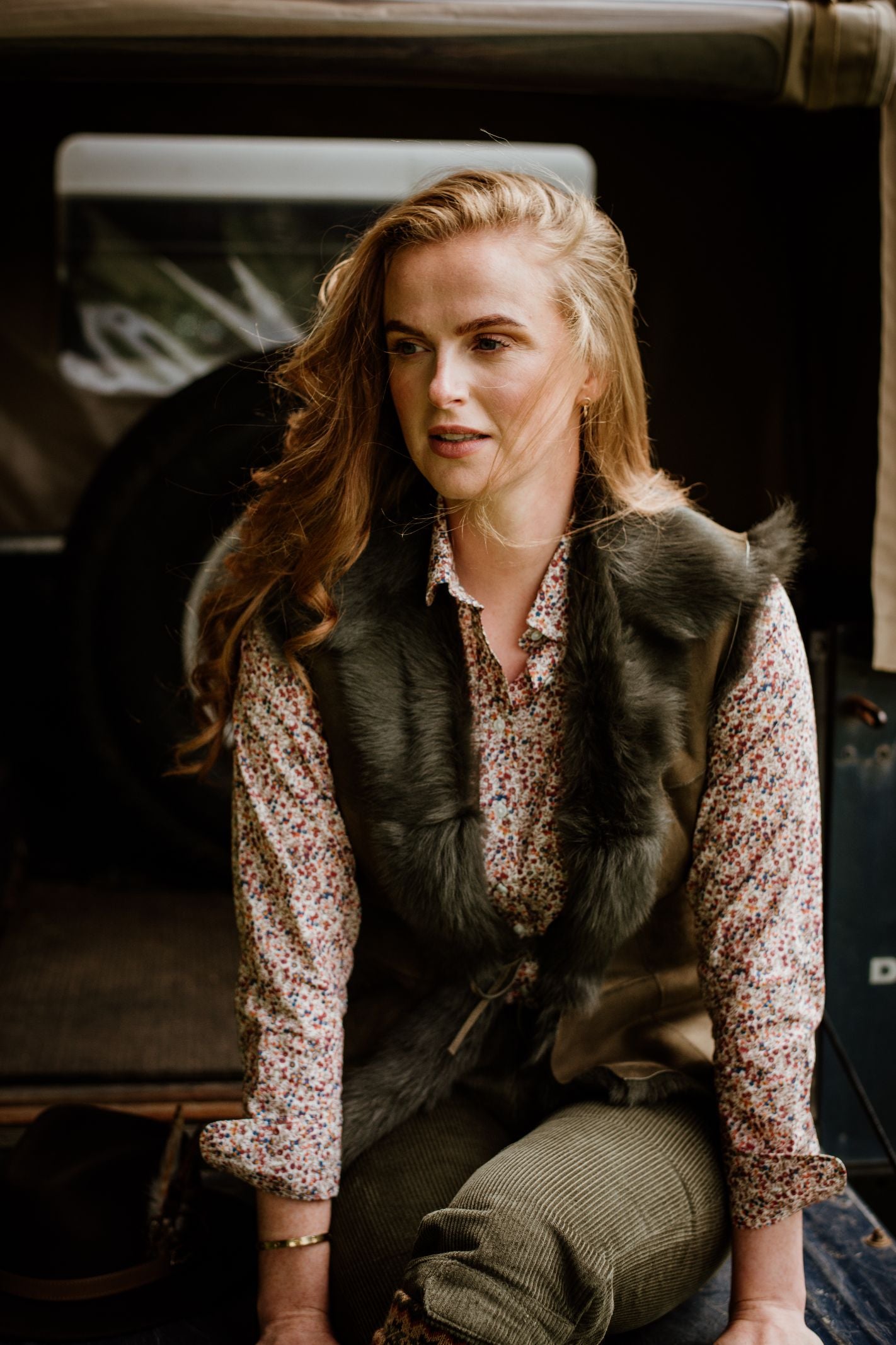 A woman with long, wavy red hair sits in a vehicle wearing a patterned shirt, corduroy pants, and the Sheepskin Reversible Gilet by Campbells of Beauly. She gazes thoughtfully to the side.