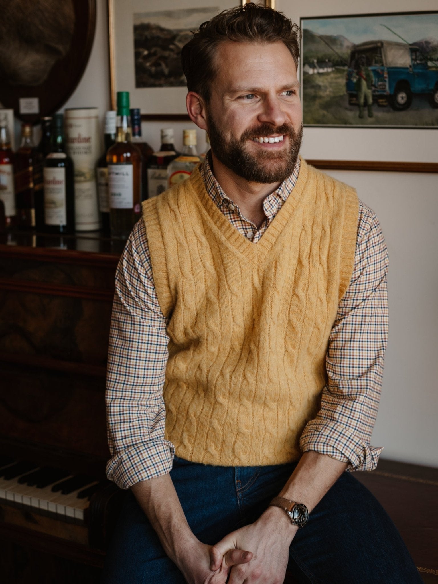 A bearded man, smiling in front of a piano with bottles and framed pictures behind him, wears the yellow Shetland Cable Slipover by Campbell's of Beauly over a plaid shirt.