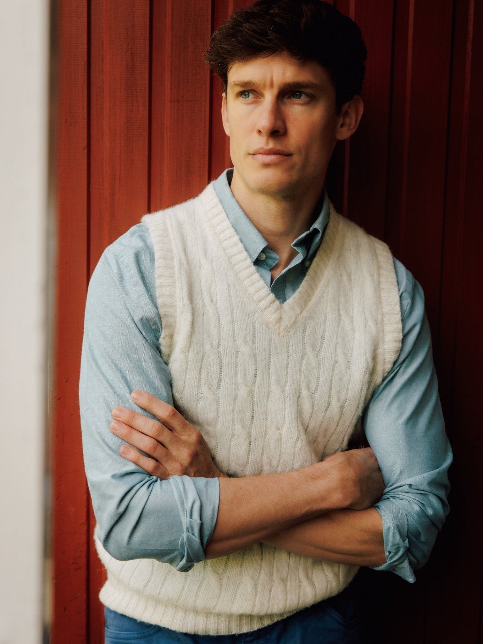A man with short brown hair, wearing a Campbell's of Beauly Oxford Button Down Shirt and a white cable-knit sweater vest, stands with arms crossed before a red wooden wall, gazing thoughtfully to the side.