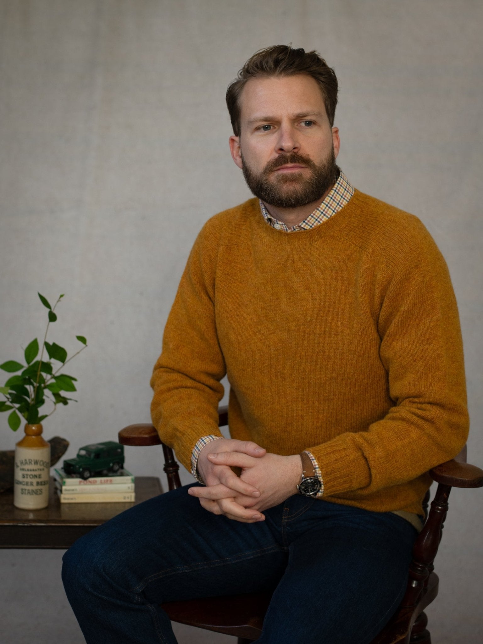 A bearded man in jeans wears a mustard Shetland Jumper by Campbell's of Beauly, sitting on a wooden chair with hands clasped. Next to him, a small table holds a plant, books, mug, and toy car against a plain background.