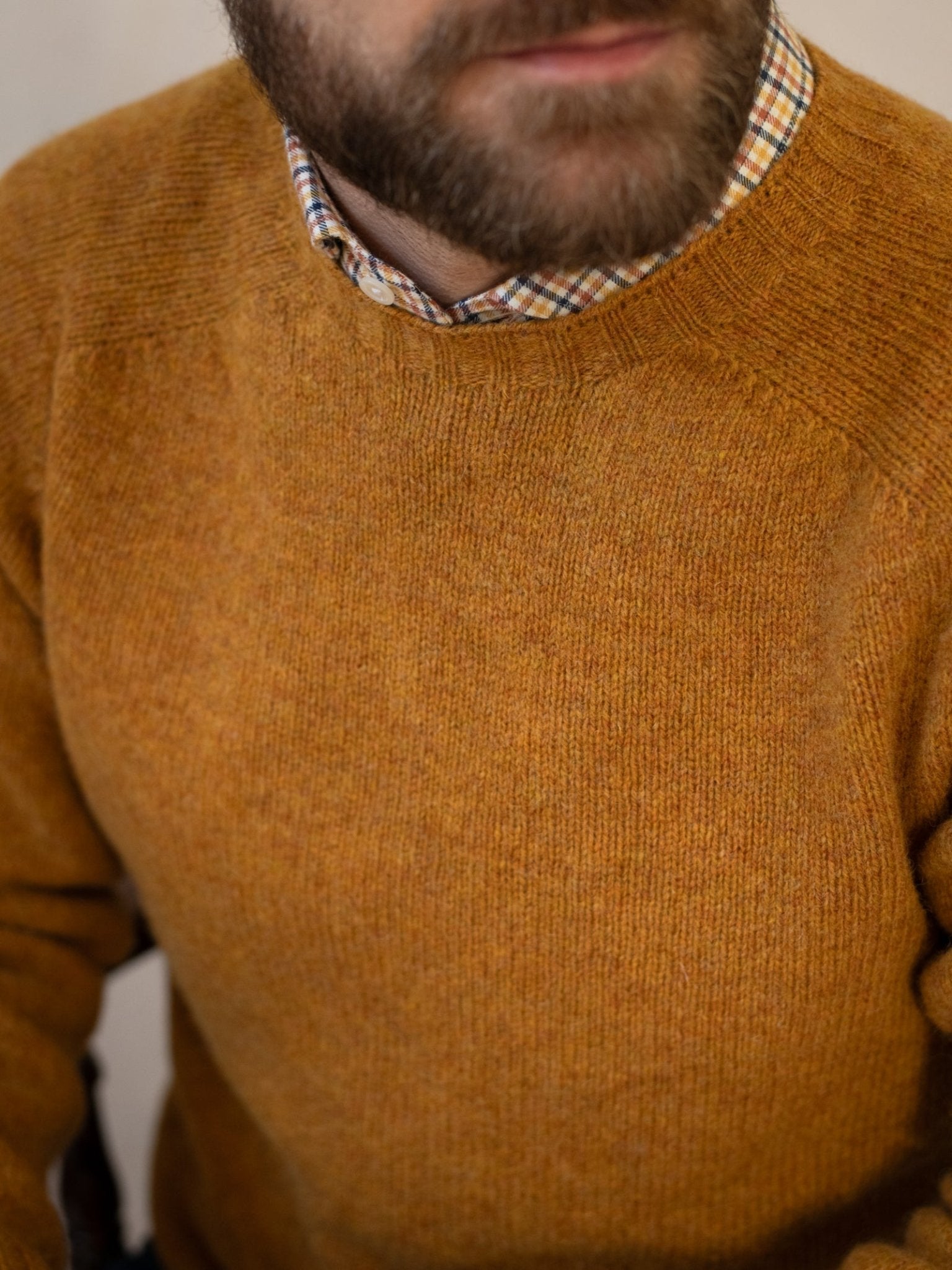 A close-up shows a bearded man wearing a Campbell's of Beauly Shetland Jumper in mustard over a plaid shirt, with his mouth and chin visible, highlighting the soft texture of Shetland lambswool.