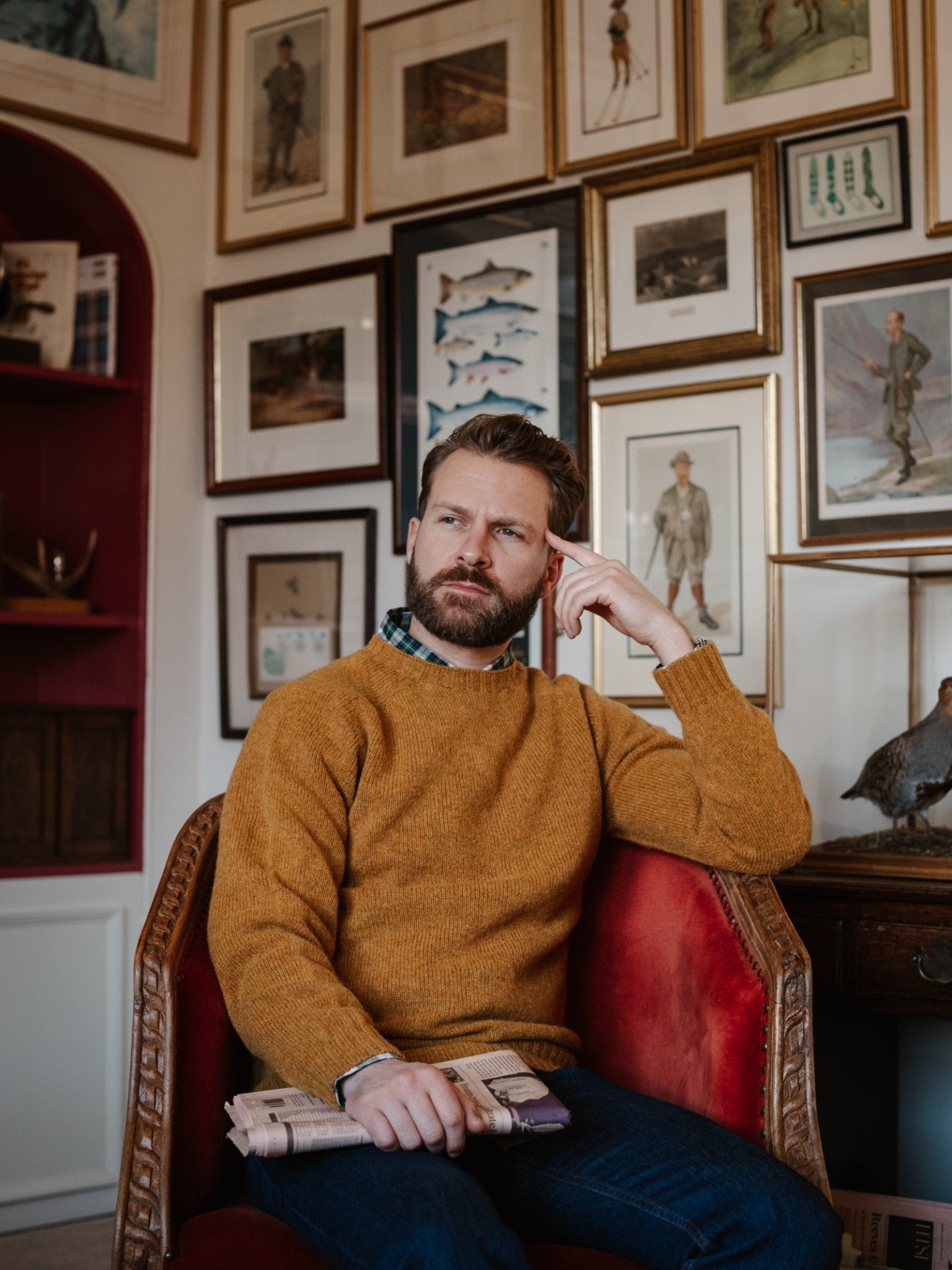 A bearded man sits thoughtfully in a red armchair, wearing a mustard Shetland Jumper by Campbell's of Beauly and holding a folded newspaper. Behind him, the wall displays framed art of fish and vintage figures.