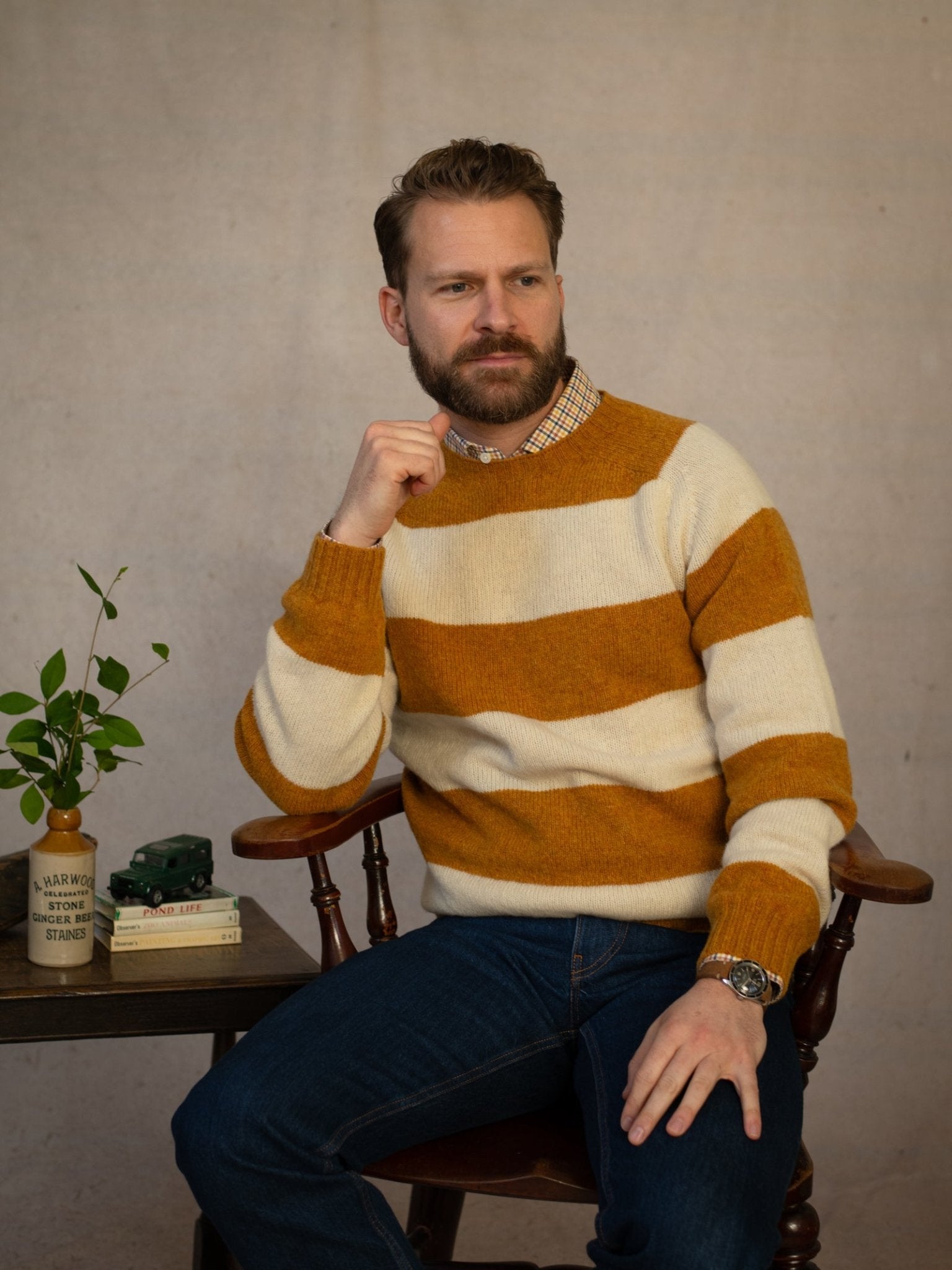 A bearded man in jeans sits thoughtfully on a wooden chair beside a small table with a plant, books, and toy car. He wears the Campbell's of Beauly Shetland Stripe Crew Jumper in Butterscotch. The background is plain and neutral.