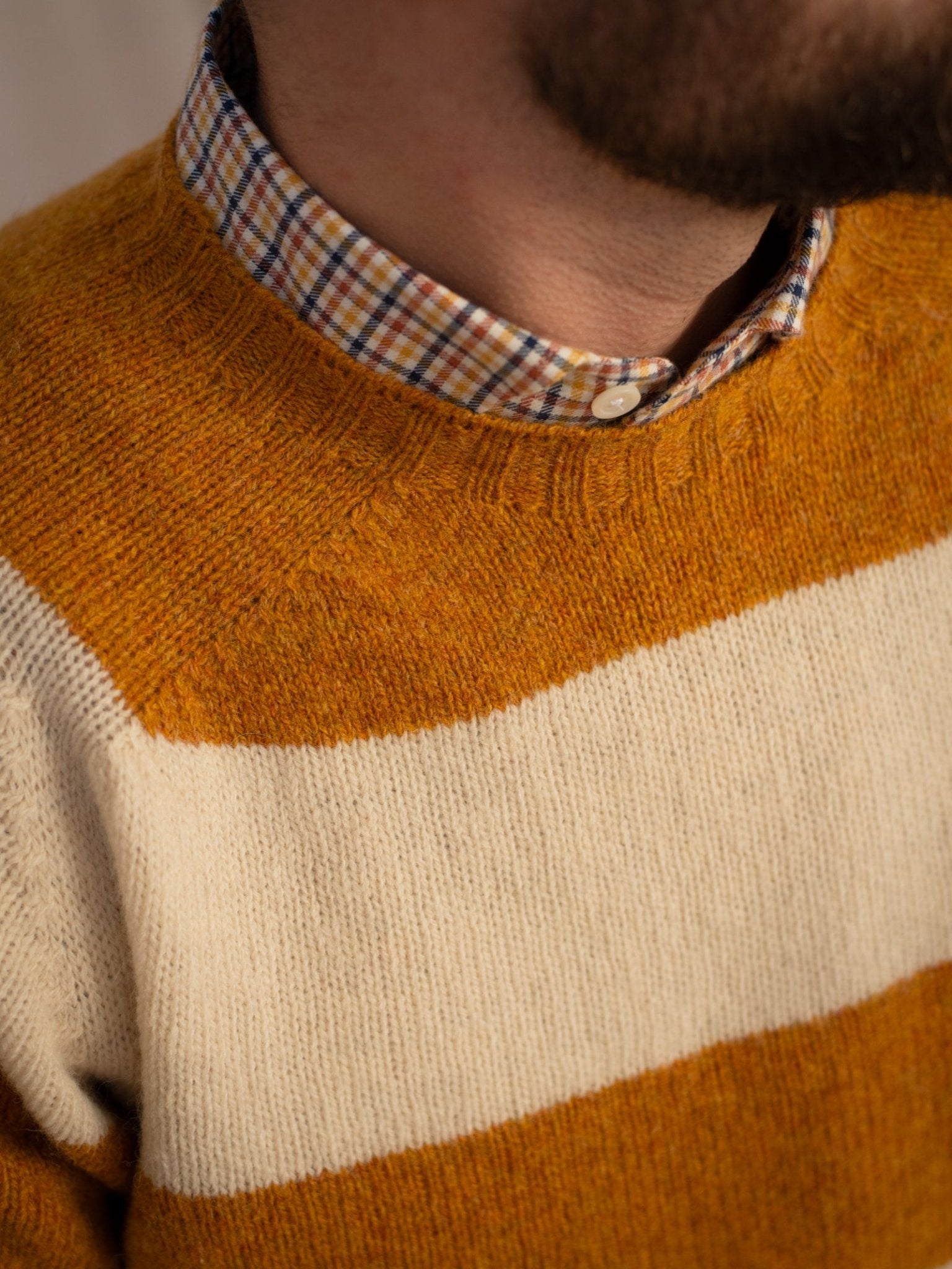 Close-up of a person in the Campbell's of Beauly Shetland Stripe Crew Jumper, featuring mustard and cream stripes, worn over a plaid shirt. Part of their neck and jaw with a short beard is also visible.