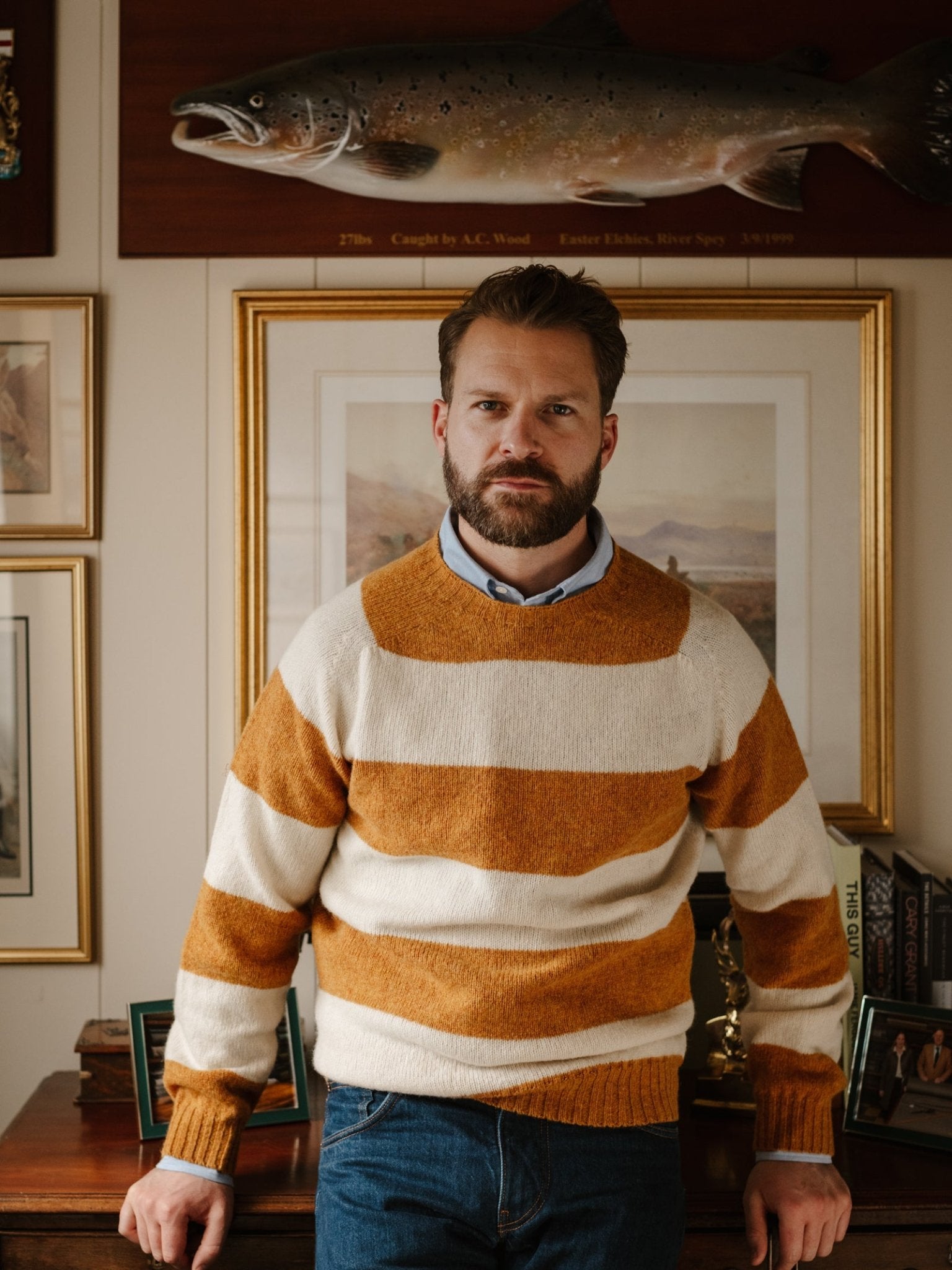 A bearded man wearing a mustard and white Campbell's of Beauly Shetland Stripe Crew Jumper stands before a wall of framed pictures and a mounted fish, with his hands resting on a wooden desk.