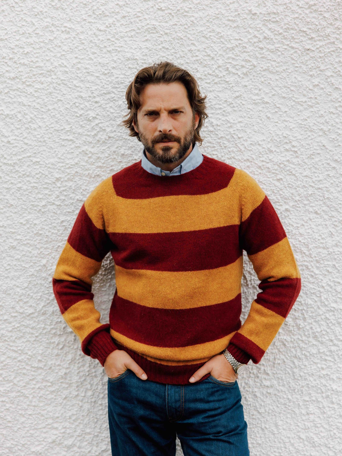 A man wearing a Campbell’s of Beauly Shetland Stripe Crew Jumper with wide yellow and maroon horizontal stripes over a light blue collared shirt.