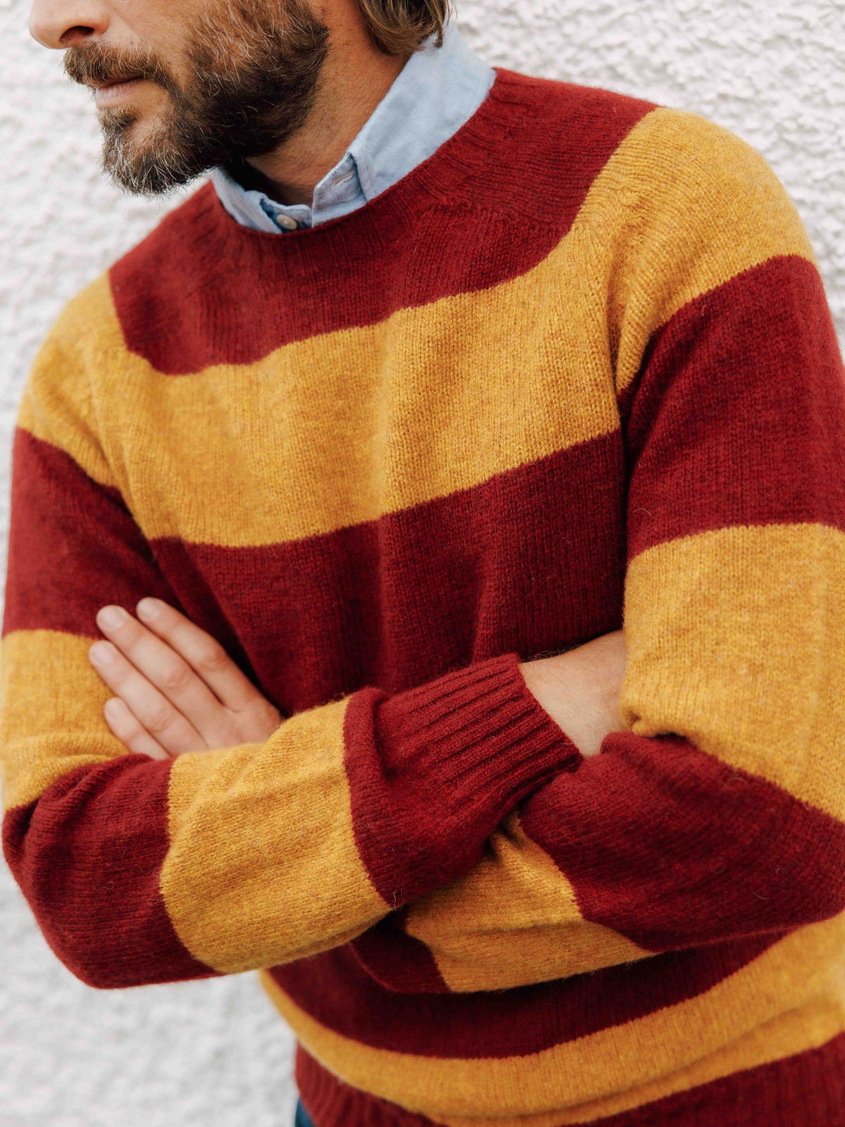A person with arms crossed, wearing a Campbell’s of Beauly Shetland Stripe Crew Jumper in wide horizontal stripes of mustard yellow and maroon over a light blue collared shirt.
