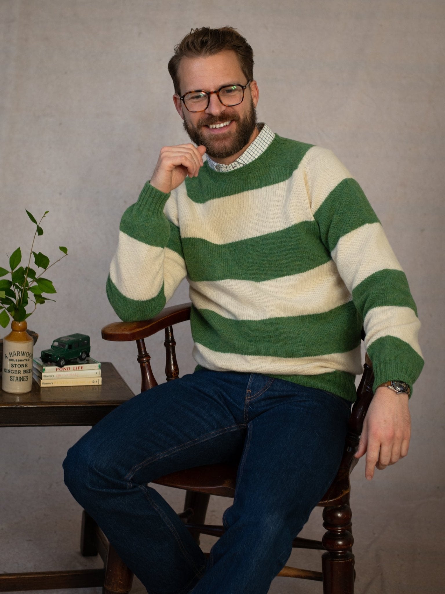 A man with glasses and a beard, wearing the Campbell's of Beauly Shetland Stripe Crew Jumper in Fern, sits on a wooden chair by a small table with books, a potted plant, and a toy car. He smiles and poses with one hand on his chin.