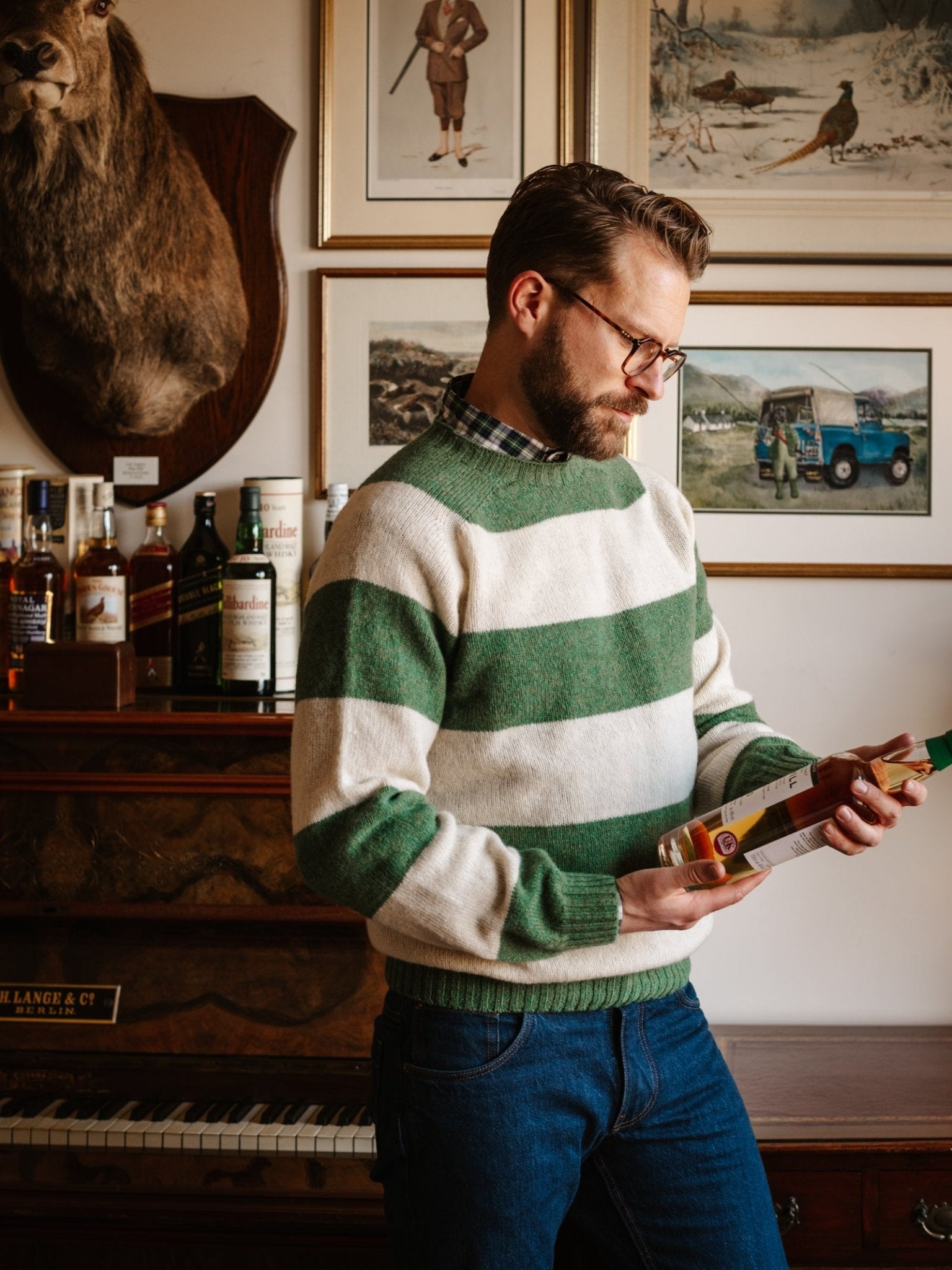 A bearded man with glasses, wearing the Campbell's of Beauly Shetland Stripe Crew Jumper and jeans, stands in a room with framed art and a piano, examining a bottle of liquor. Several bottles are displayed on the piano beside him.
