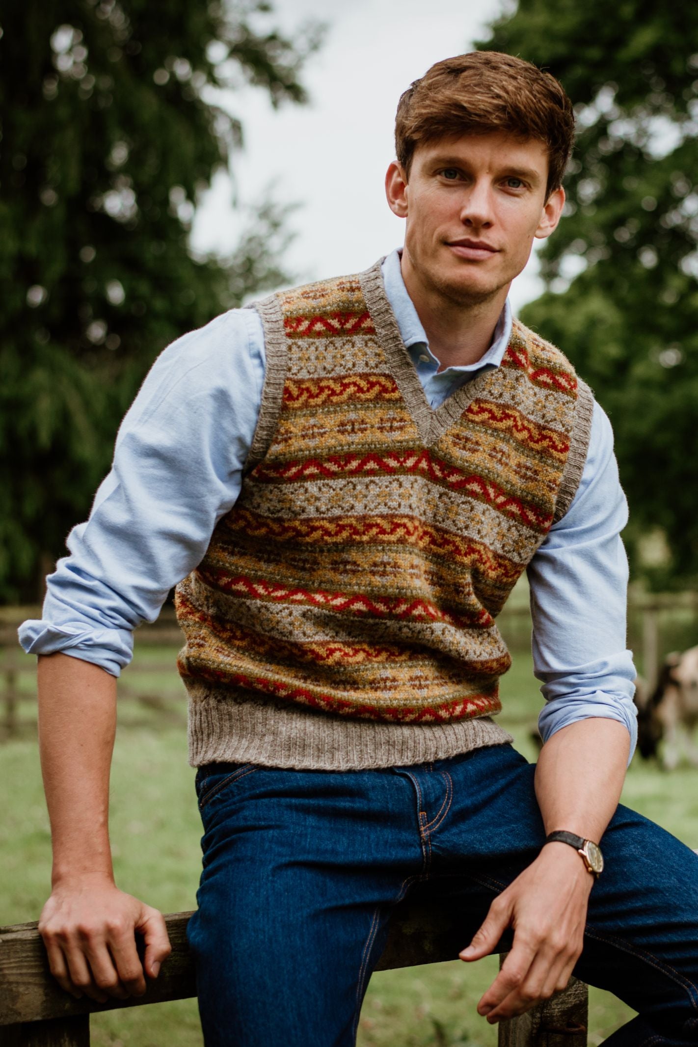 A man with short brown hair sits on a wooden fence outdoors, wearing Campbells of Beauly Fairisle Slipover over a light blue shirt and jeans, with green trees and grass in the background as he looks at the camera.