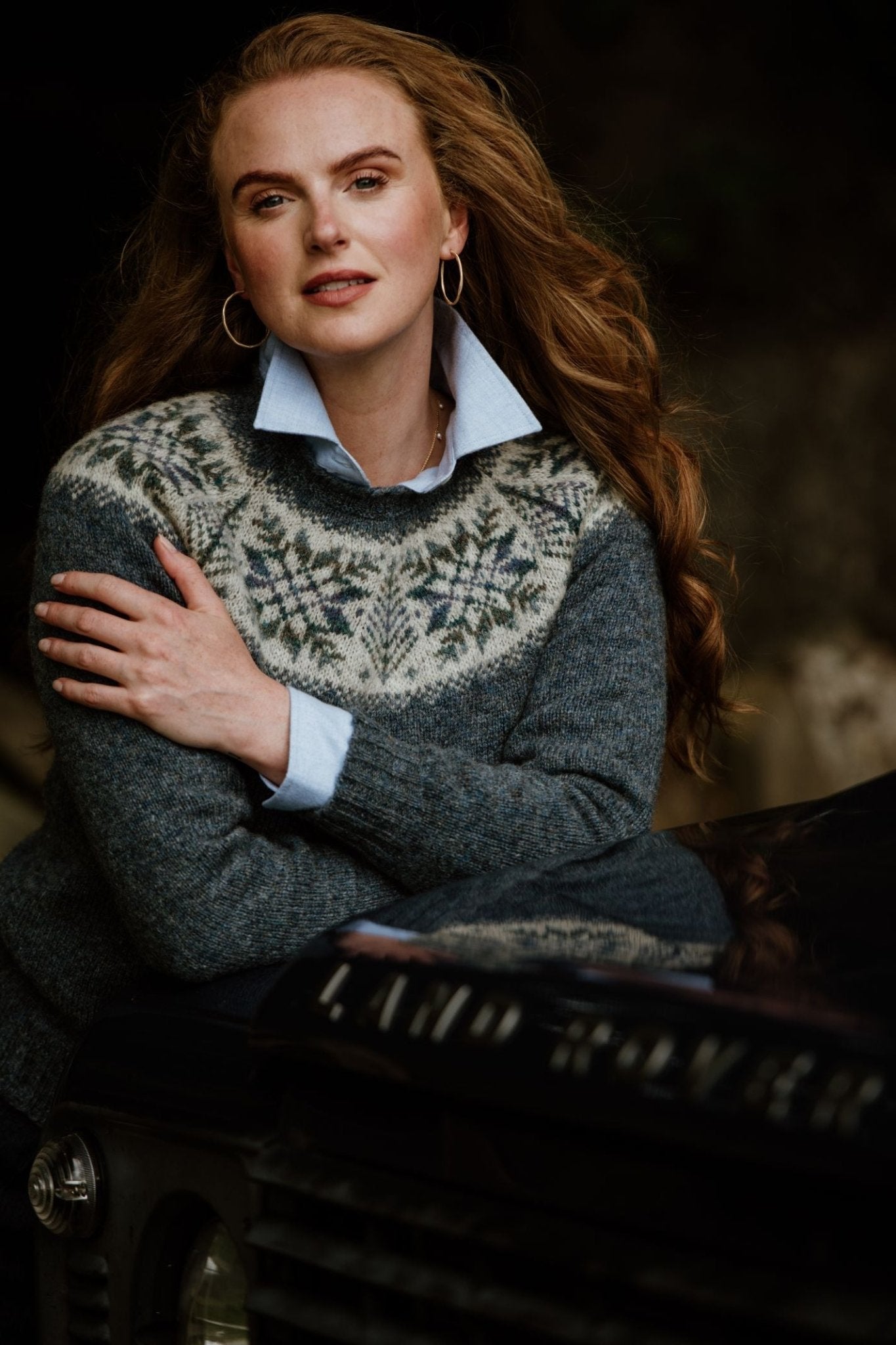 A woman with long wavy hair, wearing the Campbells of Beauly Shetland Wool Snowflake Crew Jumper over a collared shirt and gold hoop earrings, leans against a black Land Rover with its logo visible on the hood.