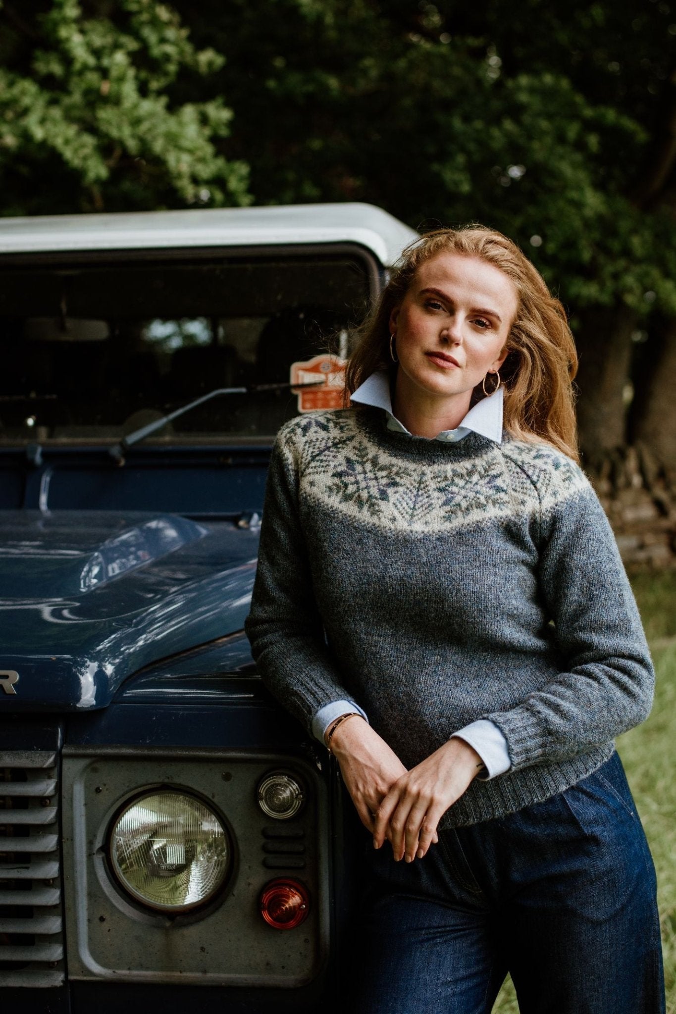 A woman with long auburn hair in blue jeans and a collared shirt leans against a dark blue vintage car, surrounded by greenery, wearing the Campbells of Beauly Shetland Wool Snowflake Crew Jumper.
