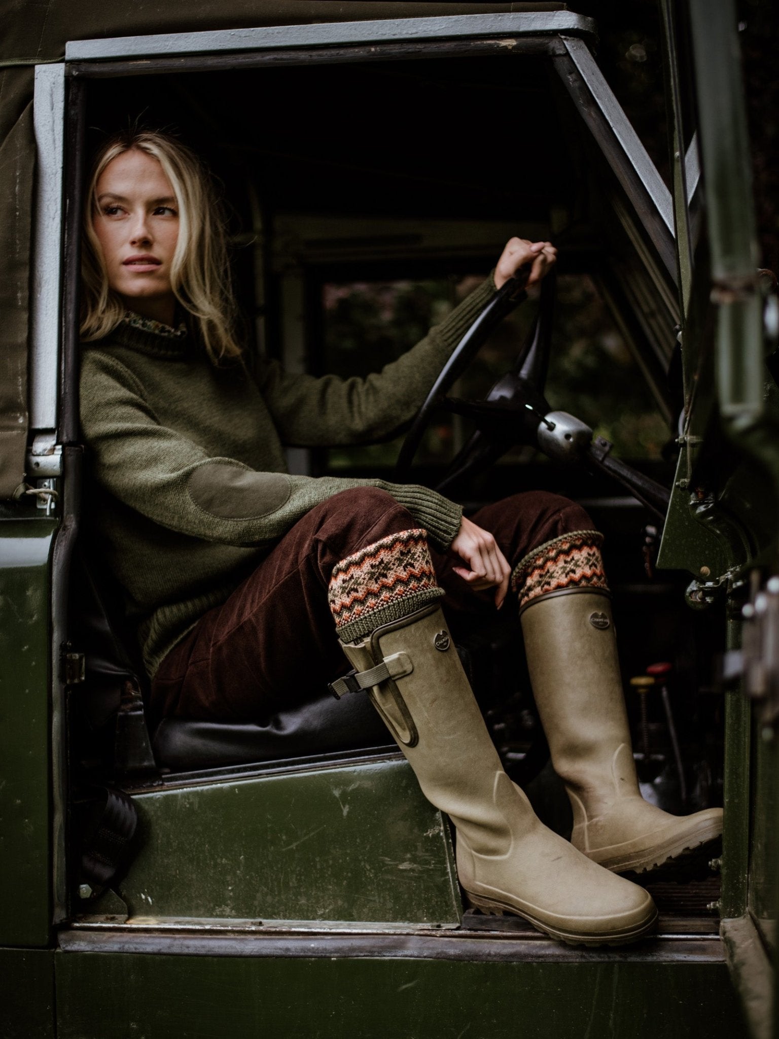 Wearing a Campbells of Beauly Crew Neck Green Shooter Jumper, a woman sits in the driver’s seat of a vintage green vehicle in the Scottish Borders, holding the wheel and looking outside.