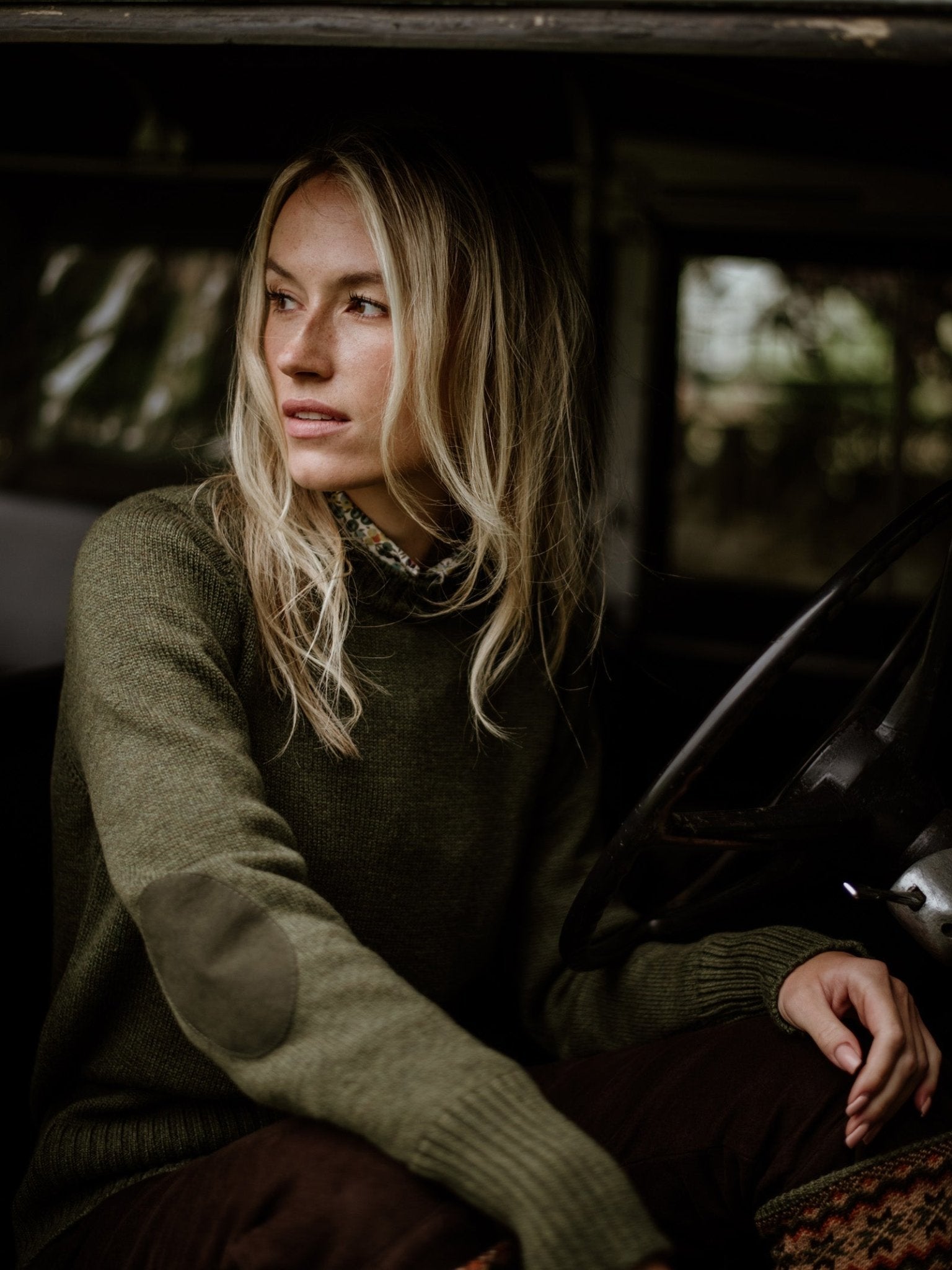 A woman with long blonde hair wears a Campbells of Beauly Crew Neck Green Shooter Jumper, sitting thoughtfully in the dimly lit drivers seat of a vehicle with the steering wheel visible.