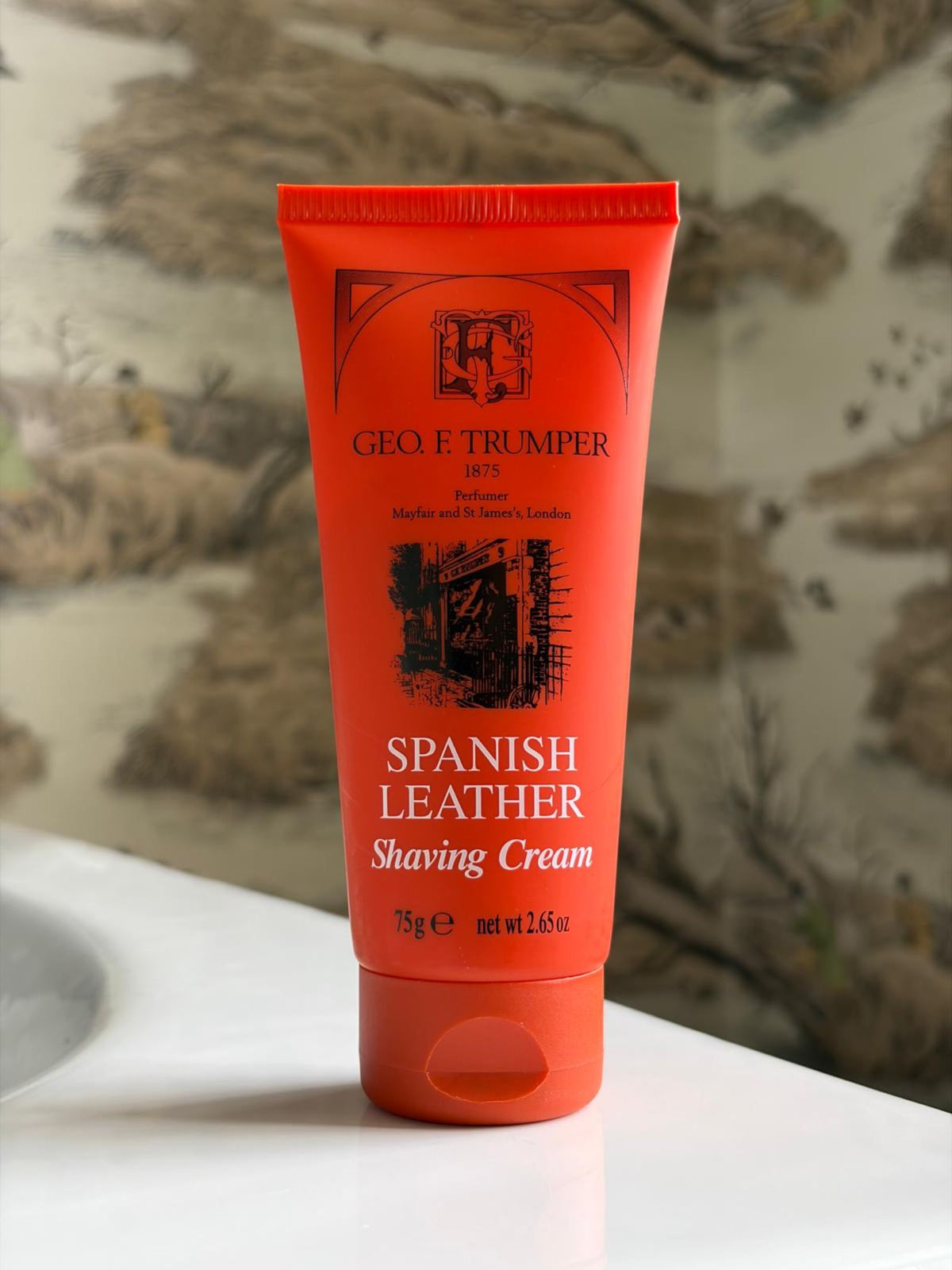 A 75g tube of Geo. F. Trumper Spanish Leather Soft Shaving Cream rests on a bathroom counter, ideal for sensitive skin, with a blurred, nature-inspired wallpaper in the background.