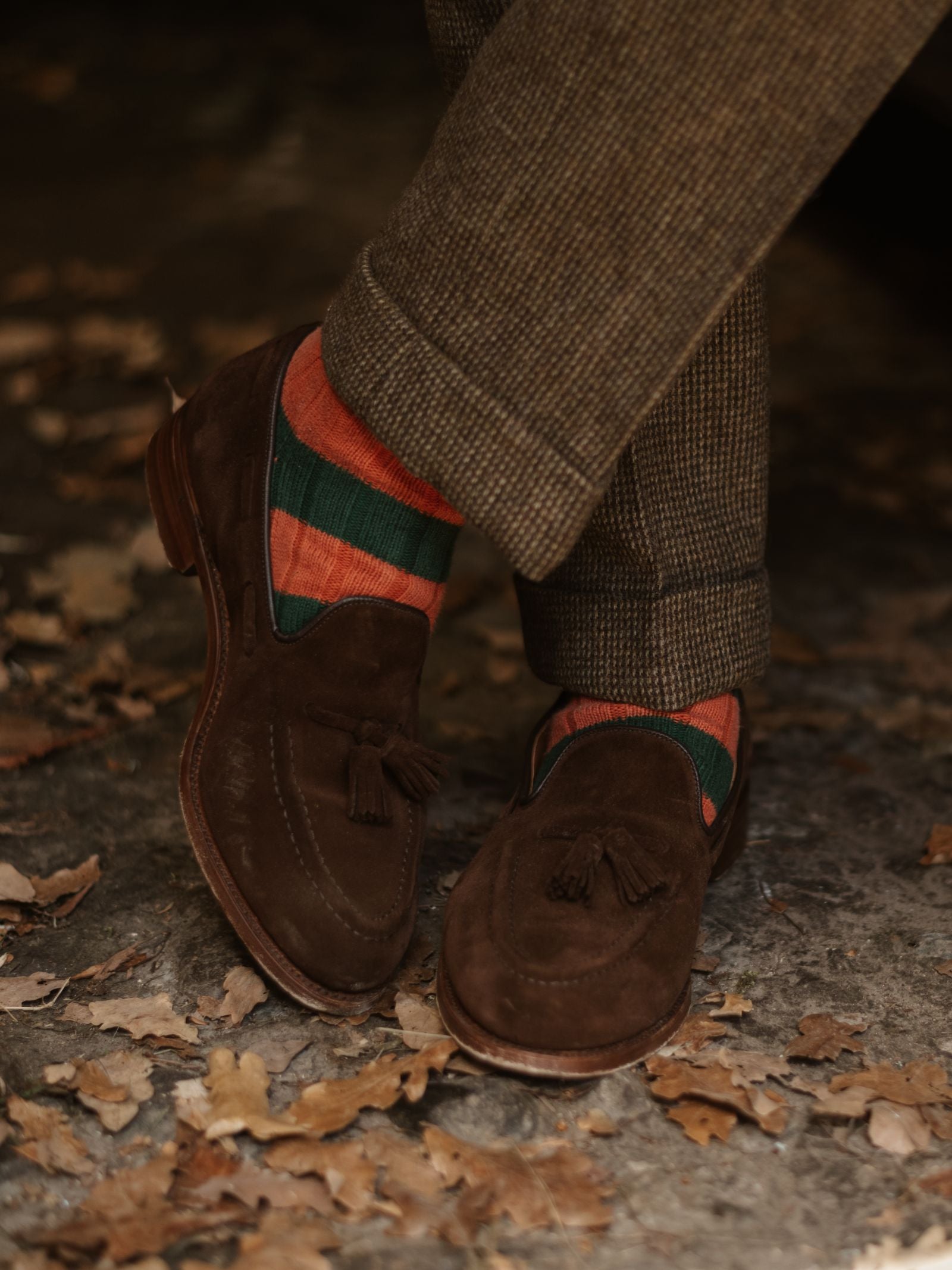 A person stands on dried leaves wearing brown suede tassel loafers, brown checkered trousers, and Campbell's of Beauly Stripe Socks.