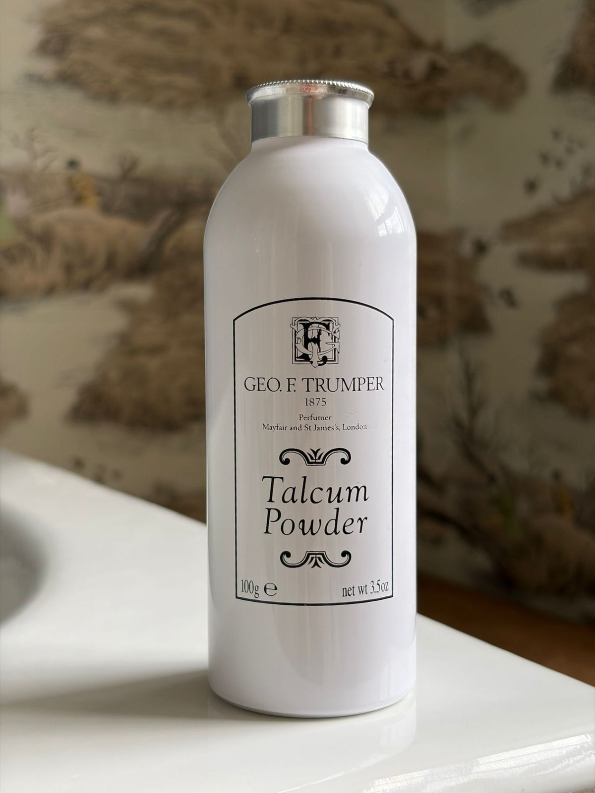 A white bottle of Geo. F. Trumper Talcum Powder, featuring cosmetic grade talc and a floral scent, rests on a bathroom counter with a blurred patterned wall behind it.