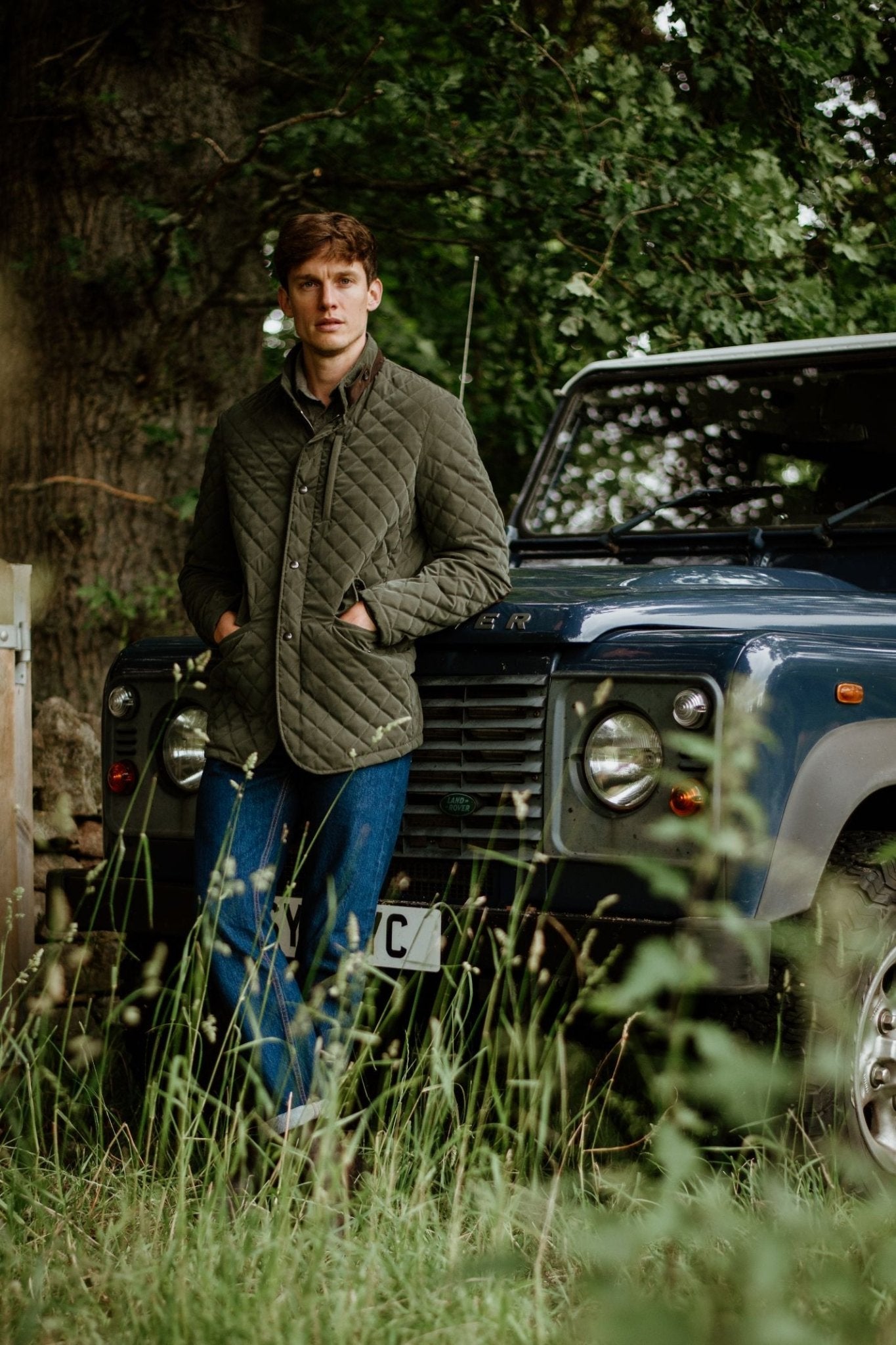 A man wears the Tomich Jacket by Campbells of Beauly, standing on grass and leaning against a dark blue off-road vehicle, with trees and greenery in the background.
