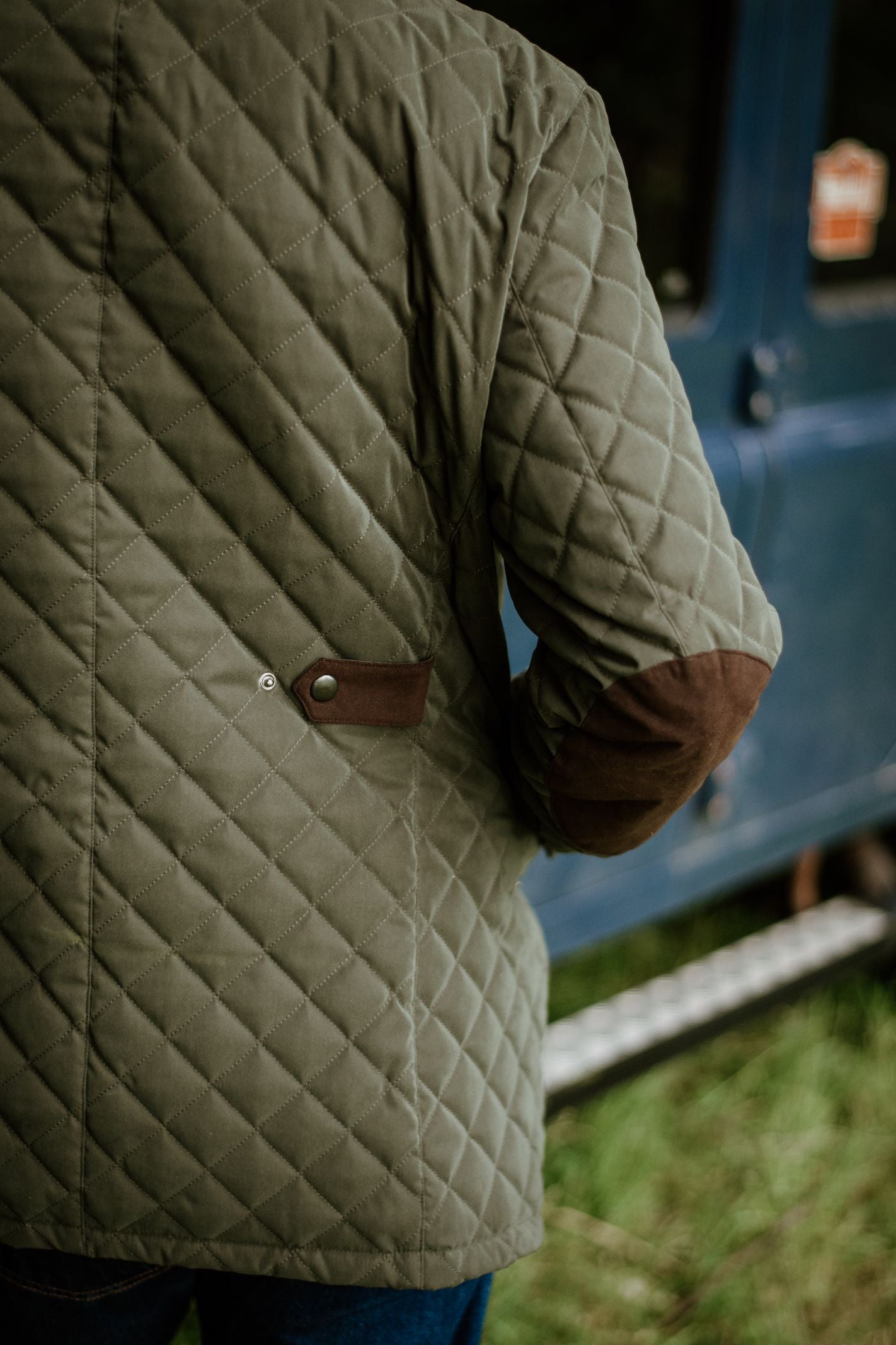 Wearing the Tomich Jacket by Campbells of Beauly, a person in classic Highland style with brown suede elbow patches stands beside a blue vehicle, highlighting the jacket’s detailed design and timeless countrywear appeal.