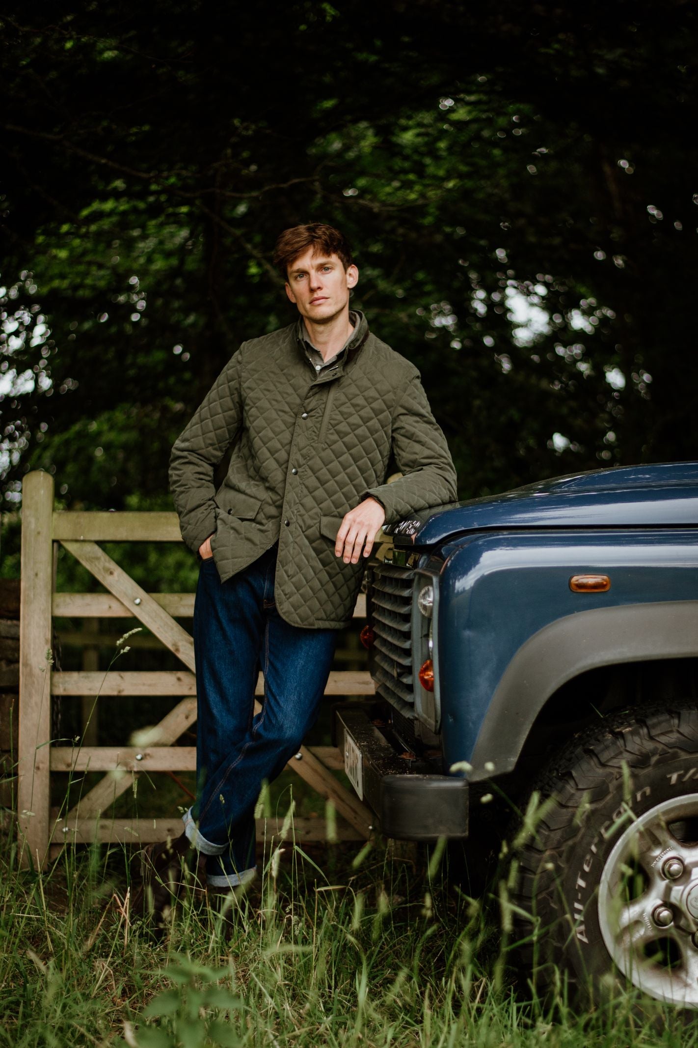 A man in Highland style wears the Campbell's of Beauly Tomich Jacket and blue jeans, leaning against a blue off-road vehicle by a wooden gate amid tall grass and trees.