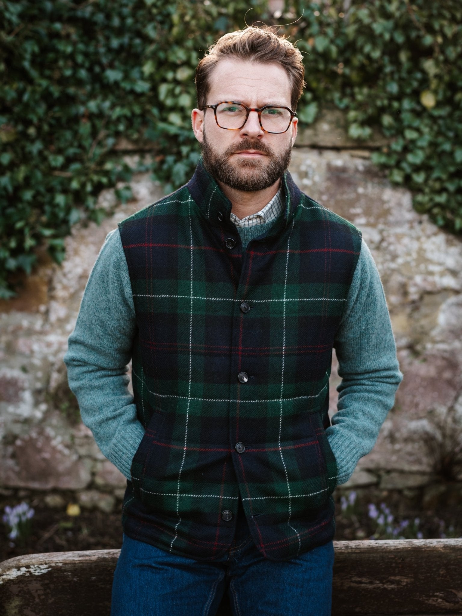 A man with glasses and a beard stands outside before an ivy-covered stone wall, wearing a Campbell's of Beauly Auld Stock Quilted Tweed Buttoned Gilet over a gray sweater and blue jeans, his hands in the gilet pockets.