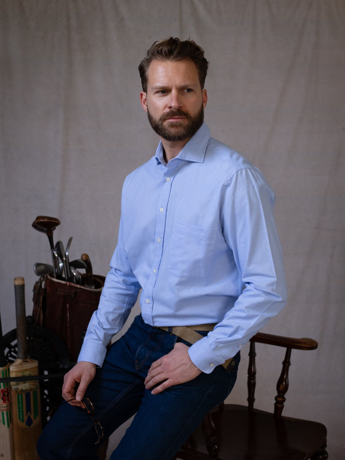 A man with a beard, dressed in the Blue Twill Shirt by Campbell’s of Beauly and jeans, sits on a wooden chair next to a set of golf clubs against a neutral background.