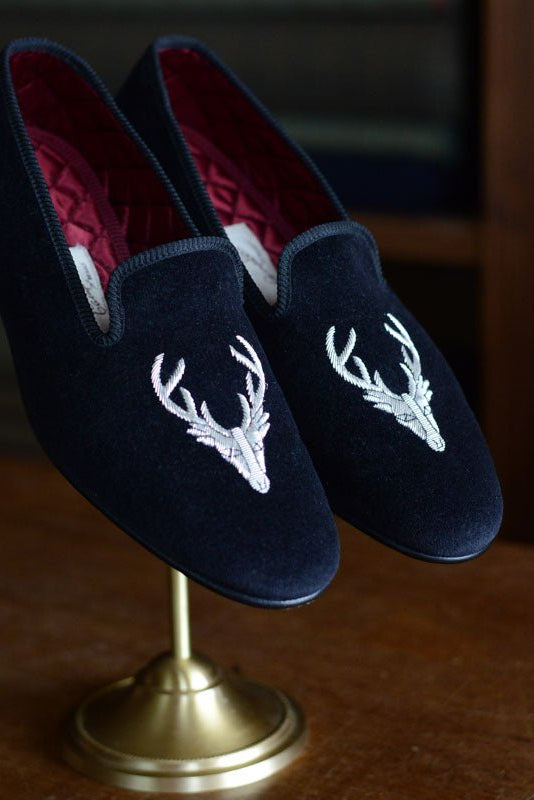 Campbells of Beauly Stag Velvet Slippers, featuring an embroidered stag motif on the toes, are ideal for evening wear and elegantly displayed on a brass stand against a wooden background.