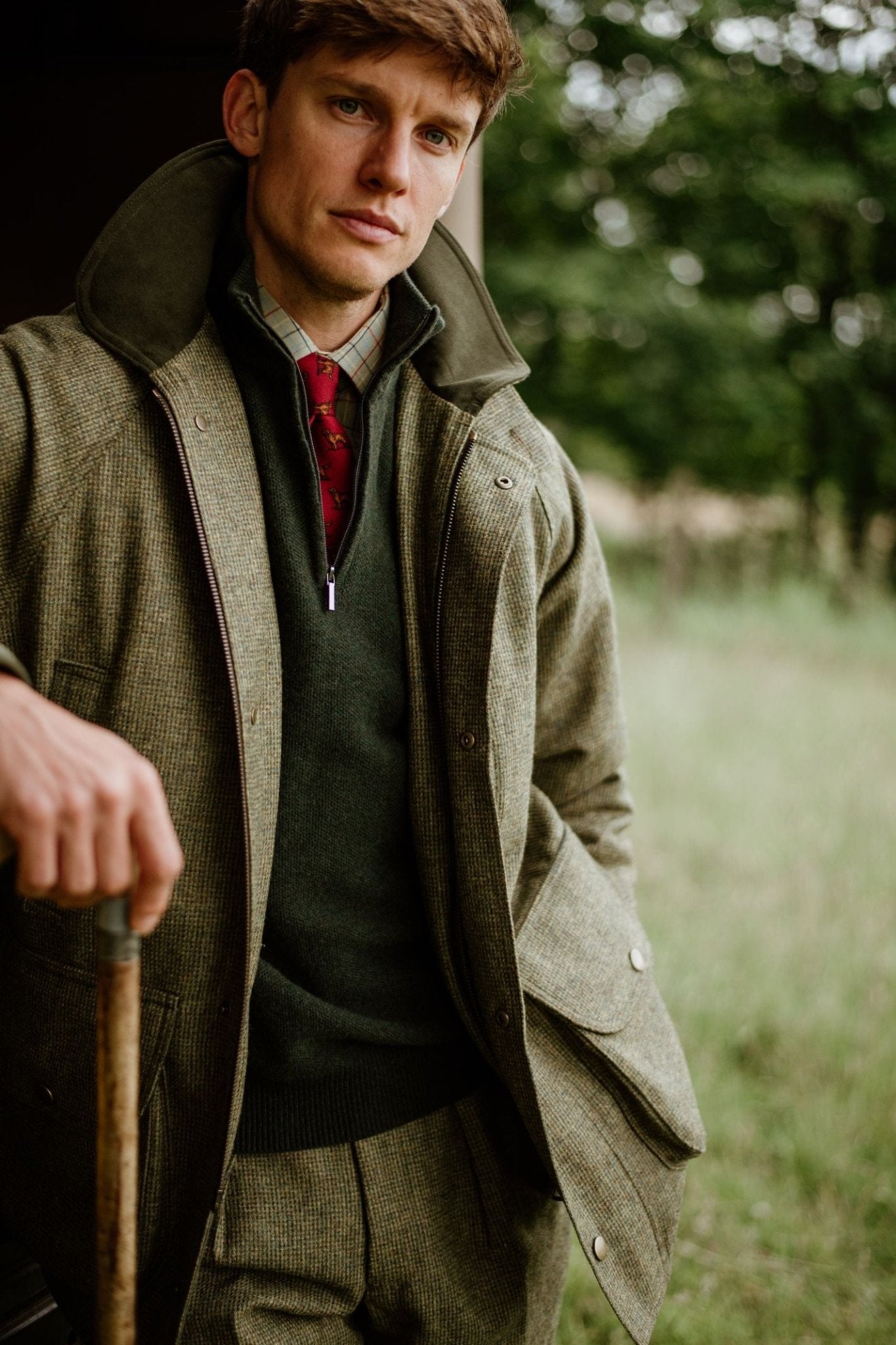 A man wearing a Campbells of Beauly Windowpane Shirt, along with a tweed jacket, vest, and trousers, plus a red tie, stands outdoors holding a walking stick against a backdrop of green grass and trees.