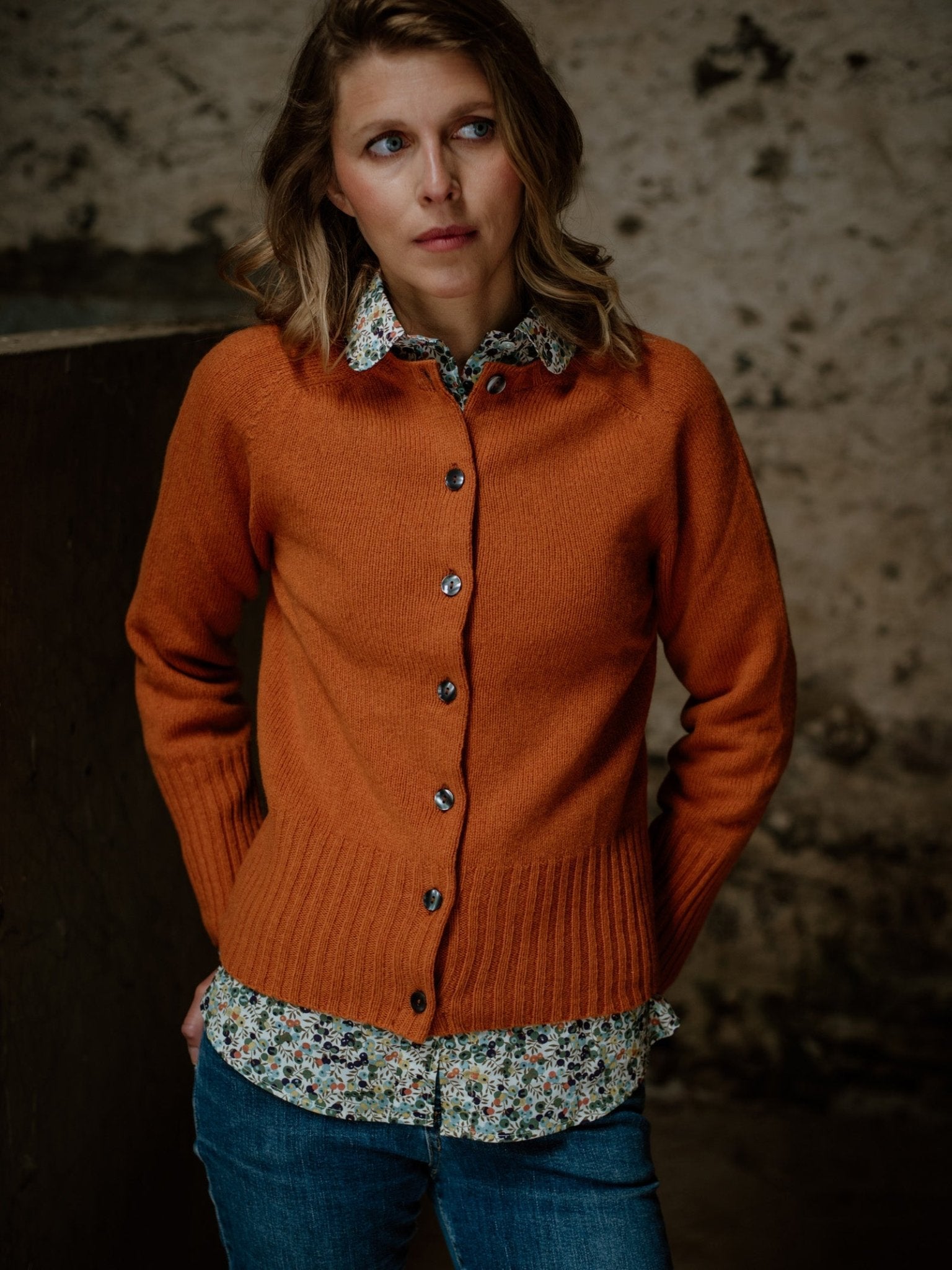 A woman with wavy blonde hair wears the Campbells of Beauly Crew Cardigan—an orange Geelong lambswool knit—over a floral shirt and blue jeans, standing against a concrete background and gazing thoughtfully to the side.
