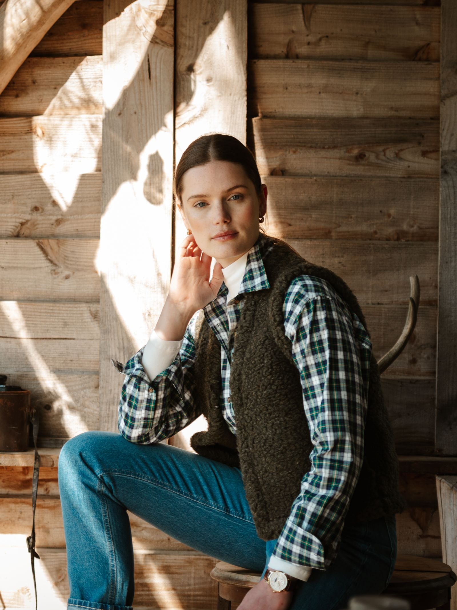 A woman wearing the Campbell's of Beauly Wool Shrug sits indoors against a rustic wooden wall, resting her chin on her hand and gazing at the camera in soft natural light.