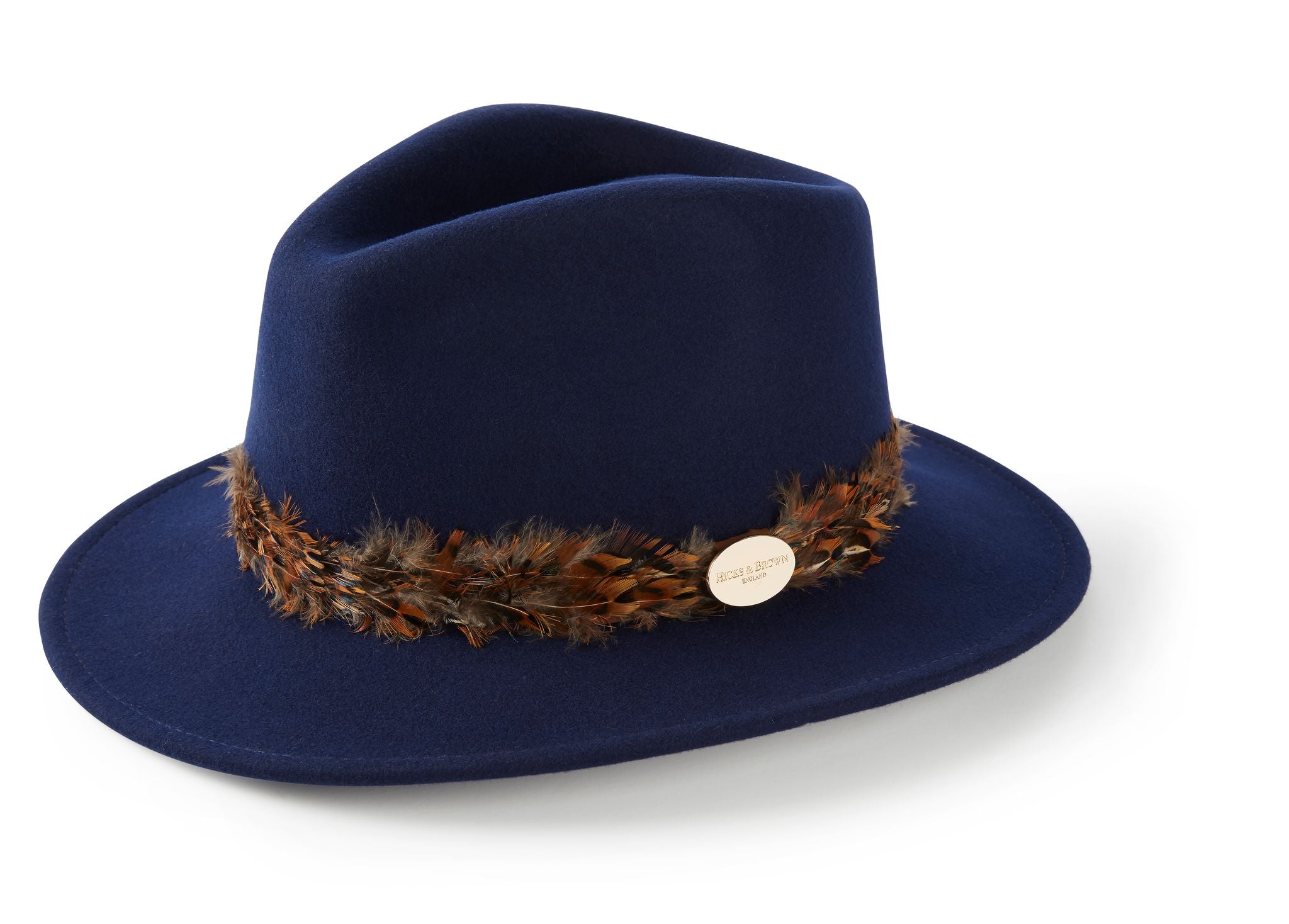 The Hicks & Brown Pheasant Feather Navy Fedora is a navy wool felt wide-brim hat, adorned with a decorative pheasant feather band and a small round gold badge on the side.