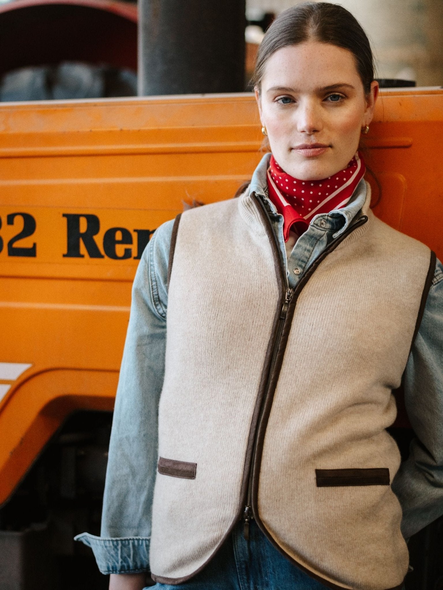 A woman wears a Campbell's of Beauly Merino Cashmere Gilet over a denim shirt and red polka dot scarf, standing before an orange vehicle with "82 Ren" visible, capturing classic countryside style.