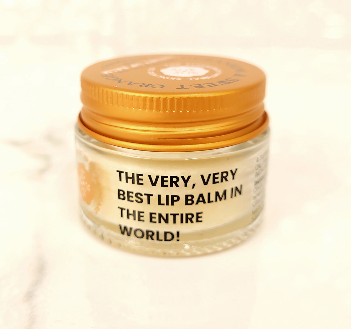 A small glass jar with an orange lid holds Geranium & Sweet Orange Luxury Lip Balm by The Edinburgh Natural Skincare Company. Bold black text on the jar reads, THE VERY, VERY BEST LIP BALM IN THE ENTIRE WORLD! Background is soft and blurred. Handmade in Scotland.