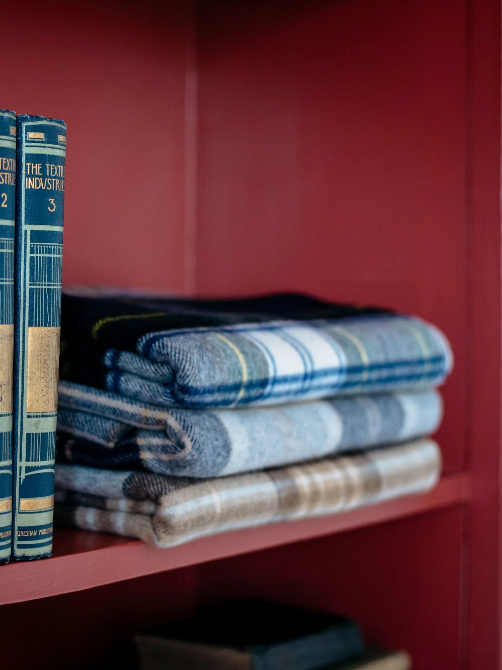 A close-up of Campbell's of Beauly Lambswool Tartan Blankets, folded and stacked on a red shelf beside vintage blue hardcover books.