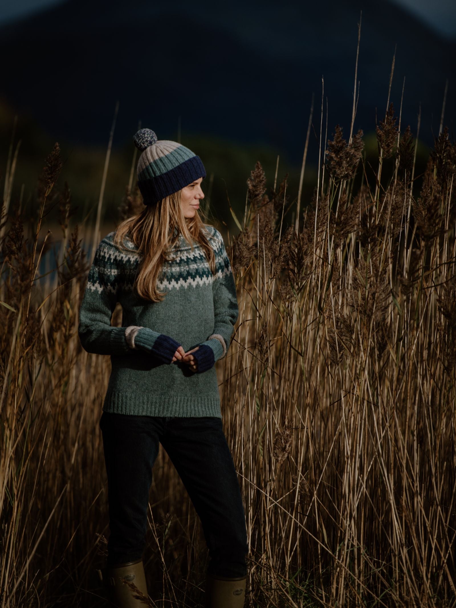 Wearing Stripe Wristwarmers by Campbells of Beauly, a long-haired woman in a patterned lambswool sweater and knitted beanie stands in tall grass, looking left toward a dark, wooded hill beneath the moody Scottish Borders sky.