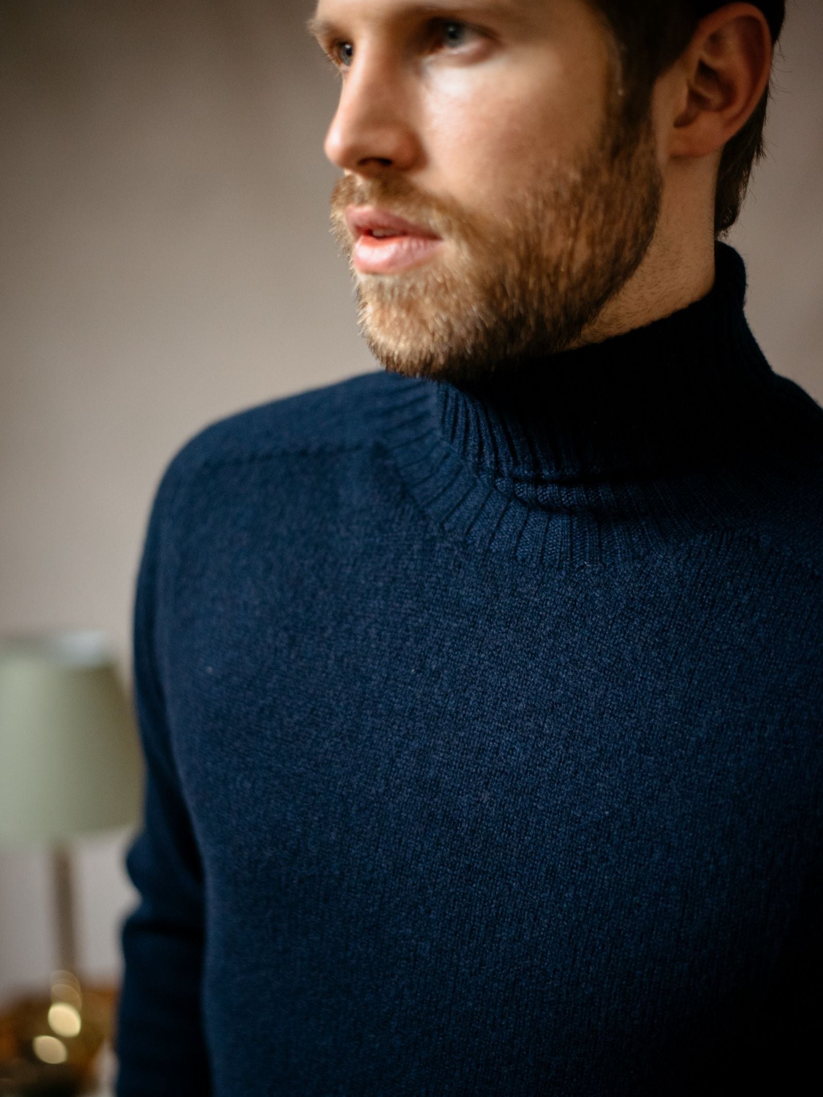 A man with a short beard wears a dark blue Campbells of Beauly Lambswool Poloneck sweater indoors. The image, slightly off-center, highlights his upper torso and face, with a blurred glowing lamp in the background for warmth.