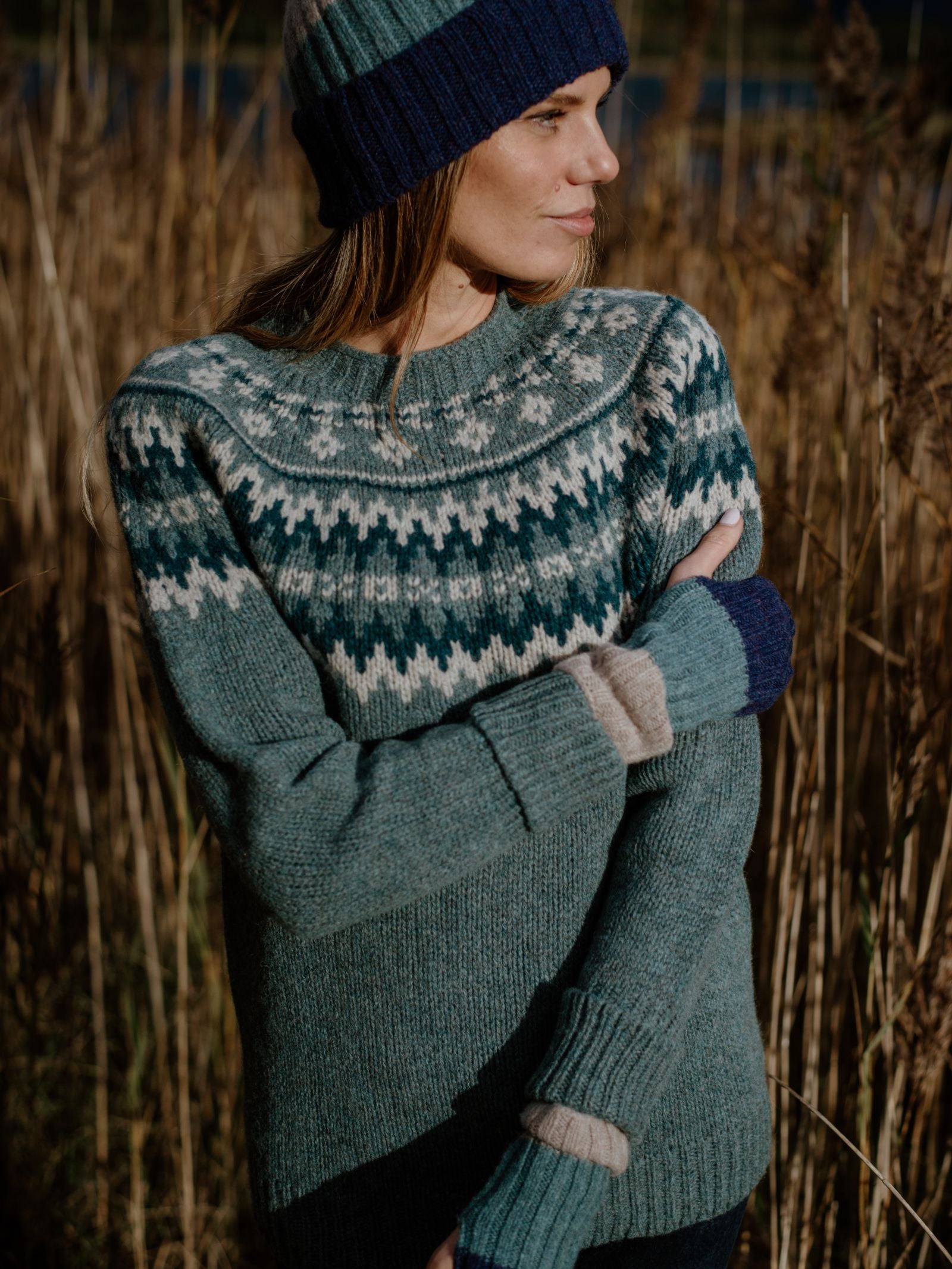 A woman stands in tall dry grass, wearing a green and cream lambswool sweater, blue-green knit hat, and Campbells of Beauly Stripe Wristwarmers. She looks to the side with her arms crossed as sunlight highlights her face and knitwear.