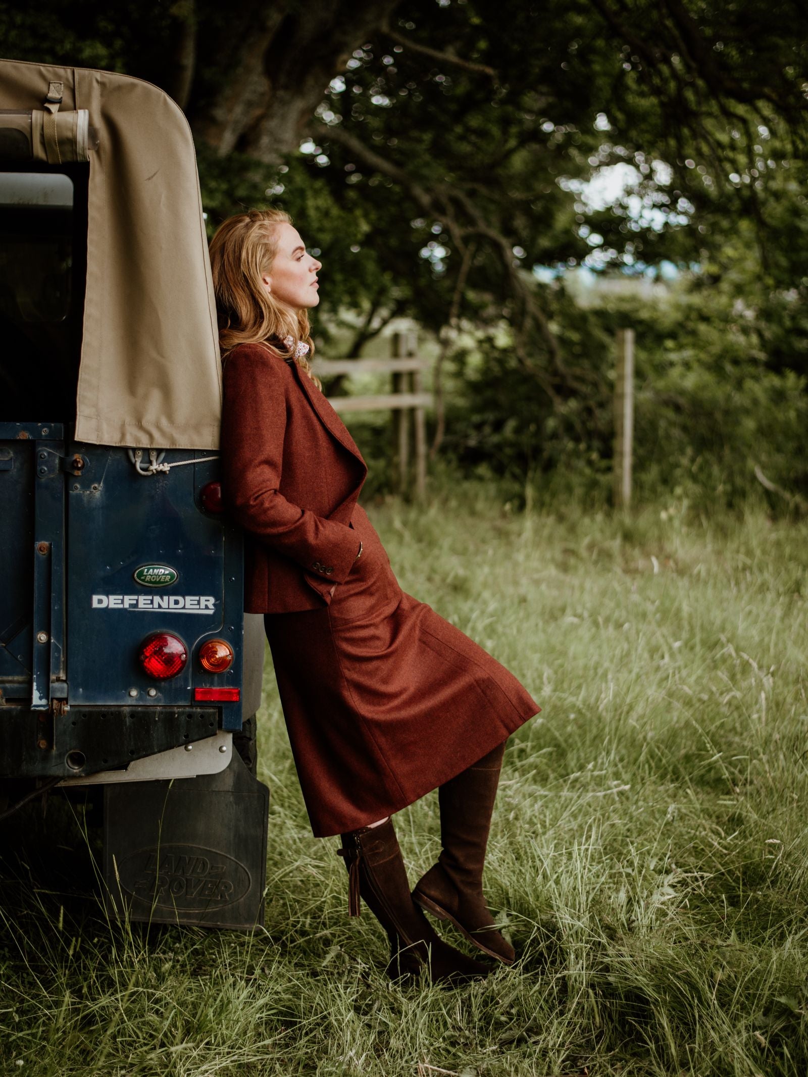 A woman wearing the House Jacket by Campbell's of Beauly and boots leans against a blue vehicle in a grassy field, with trees and a fence behind her. She looks relaxed and thoughtful, gazing off to the side.
