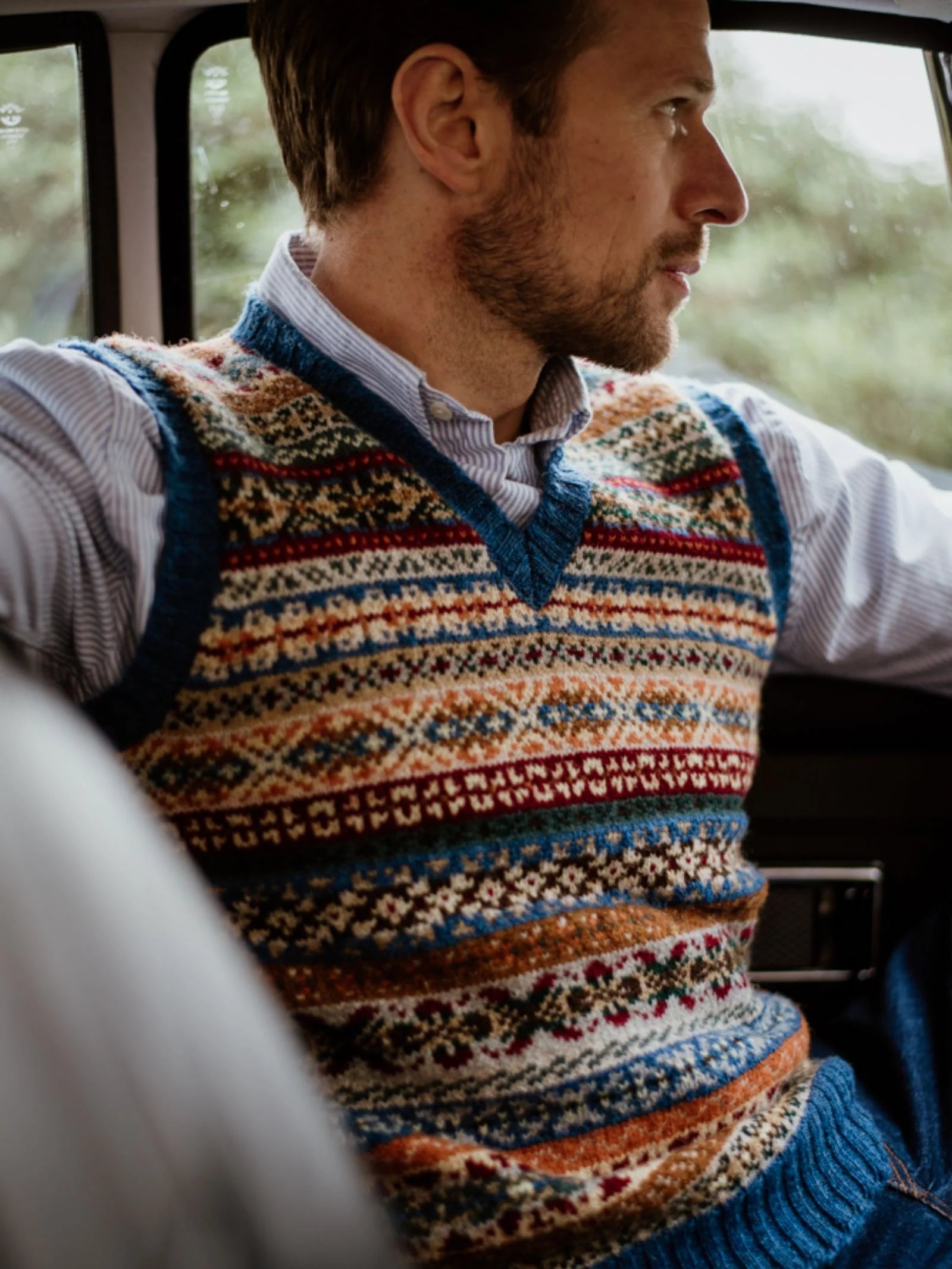 A man wearing a Campbell’s of Beauly Fairisle Slipover in a blue, orange, and cream Shetland wool pattern sits in a car, looking out the window.