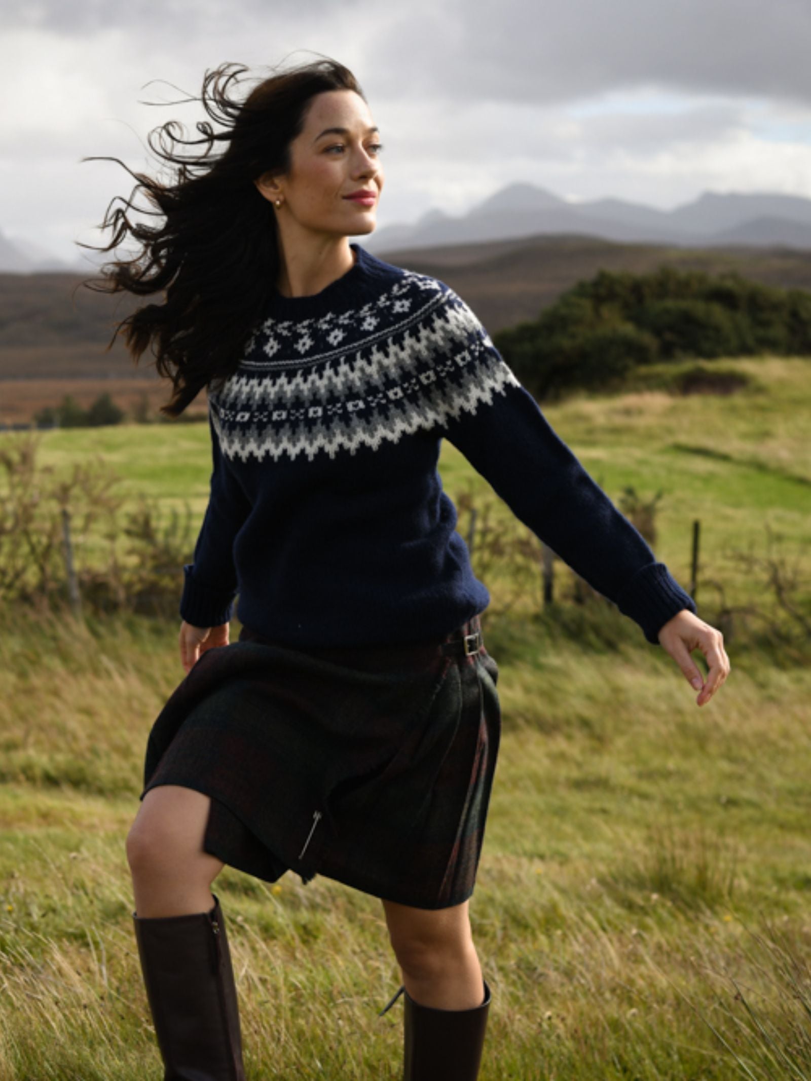 A woman with long dark hair wears the Campbell's of Beauly Chunky Fairisle Jumper, a dark plaid skirt, and brown boots as she strolls through a grassy field with hills and mountains behind her, looking content.
