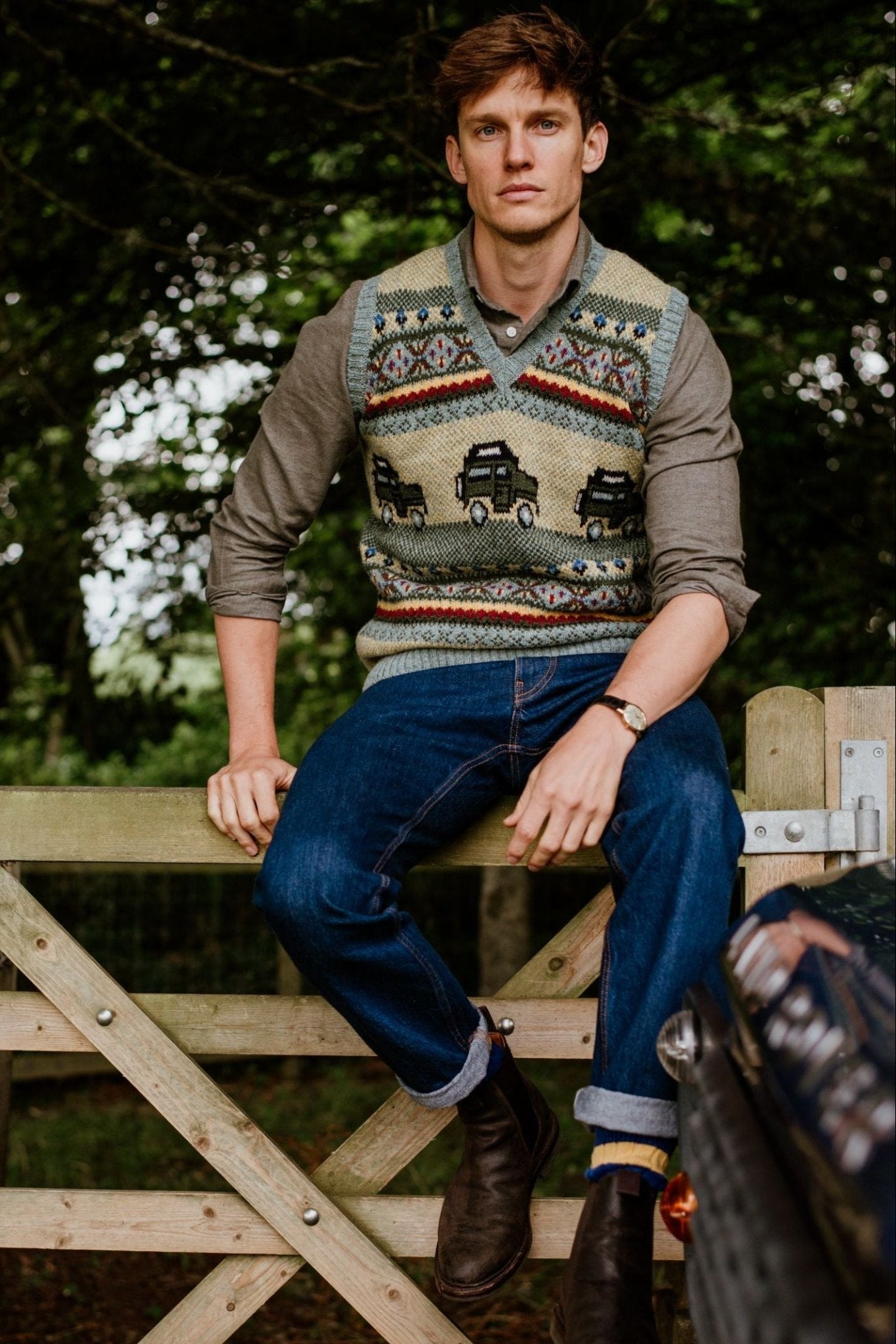 A man with short brown hair sits on a wooden gate outdoors, wearing the Campbells of Beauly Land Rover Defender Fairisle Slipover over a button-down shirt, with rolled-up jeans and brown boots amid trees and greenery.