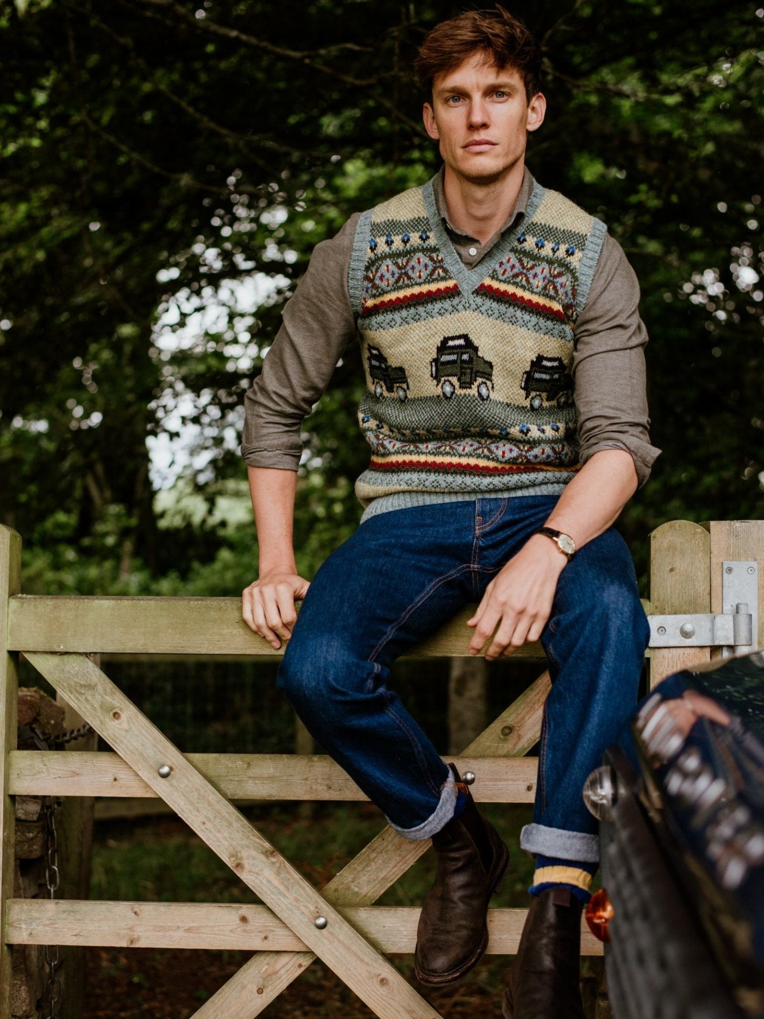 A man with short brown hair sits on a wooden gate outdoors, wearing the Campbells of Beauly Land Rover Defender Fairisle Slipover over a button-down shirt, with rolled-up jeans and brown boots amid trees and greenery.